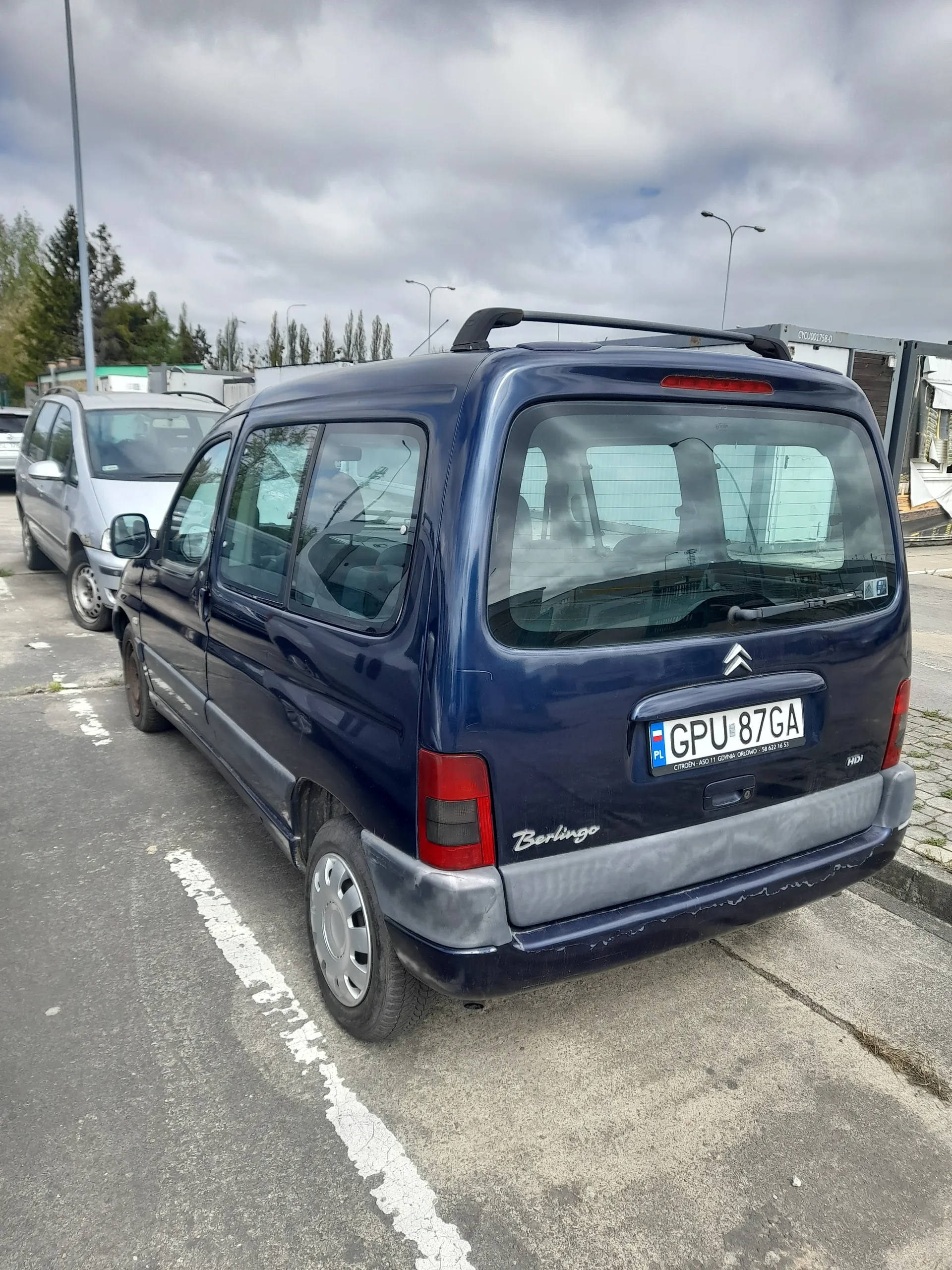 Zdjęcie ciemnoniebieskiego samochodu Citroën Berlingo zaparkowanego na asfaltowej powierzchni z białą linią. Widoczny tył pojazdu z tablicą rejestracyjną GPU 87GA, logo Citroëna, czerwonymi światłami tylnymi oraz napisem Berlingo. W tle inne samochody, drzewa, latarnie uliczne i zachmurzone niebo. Samochód znajduje się na parkingu, a obraz jest obrócony o 90 stopni w prawo.