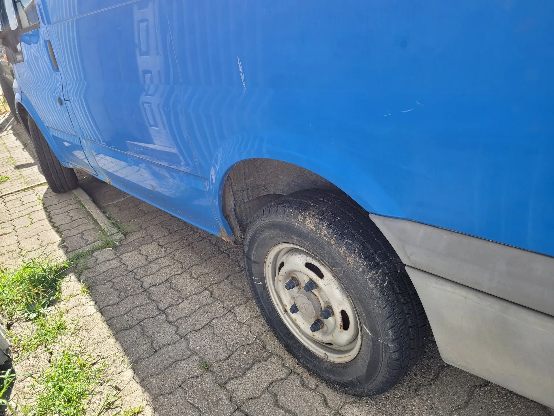 Zbliżenie na dolną część niebieskiego vana zaparkowanego na kostce brukowej. Widoczny jest tylny lewy bok pojazdu z kołem na białej stalowej feldze oraz szarym plastikowym pasem ochronnym u dołu nadwozia. Obok samochodu znajduje się krawężnik i pas zieleni z trawą.