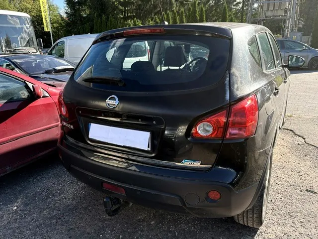 Tylny widok czarnego samochodu marki Nissan w nadwoziu hatchback, zaparkowanego na utwardzonym placu. Samochód ma widoczne logo Nissana na klapie bagażnika oraz zasłoniętą tablicę rejestracyjną. Po lewej stronie znajduje się fragment czerwonego samochodu, a w tle biały furgon i inne pojazdy oraz zielone drzewa.