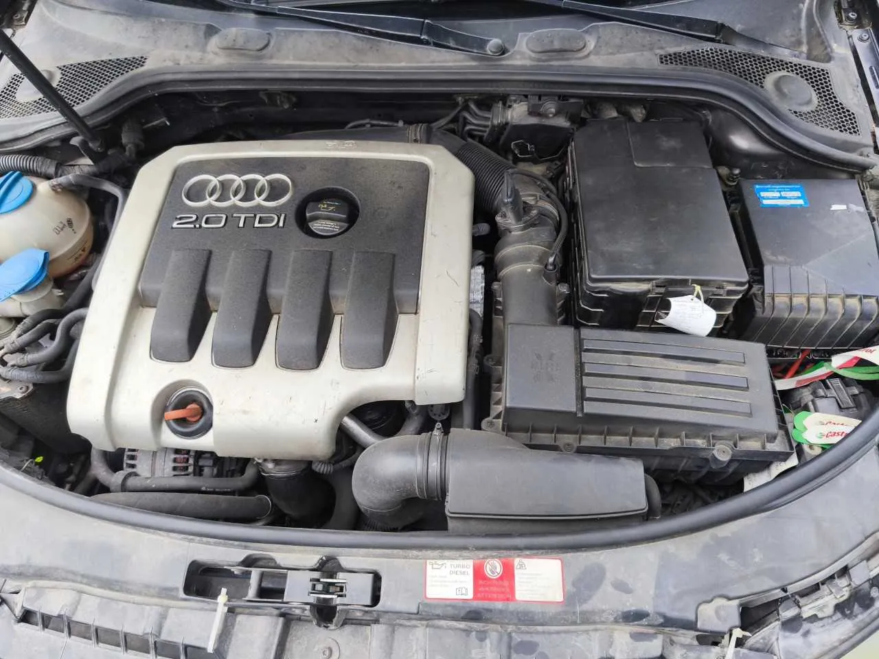 Widok z góry na komorę silnika samochodu. W centrum znajduje się srebrna osłona silnika z logo Audi oraz napisem '2.0 TDI'. Po prawej stronie widoczne są czarne skrzynki z elektroniką i akumulator. Poniżej osłony silnika znajduje się czarny wlot powietrza. Na dolnej krawędzi maski widoczna jest czerwona naklejka ostrzegawcza dotycząca silnika turbo diesel.