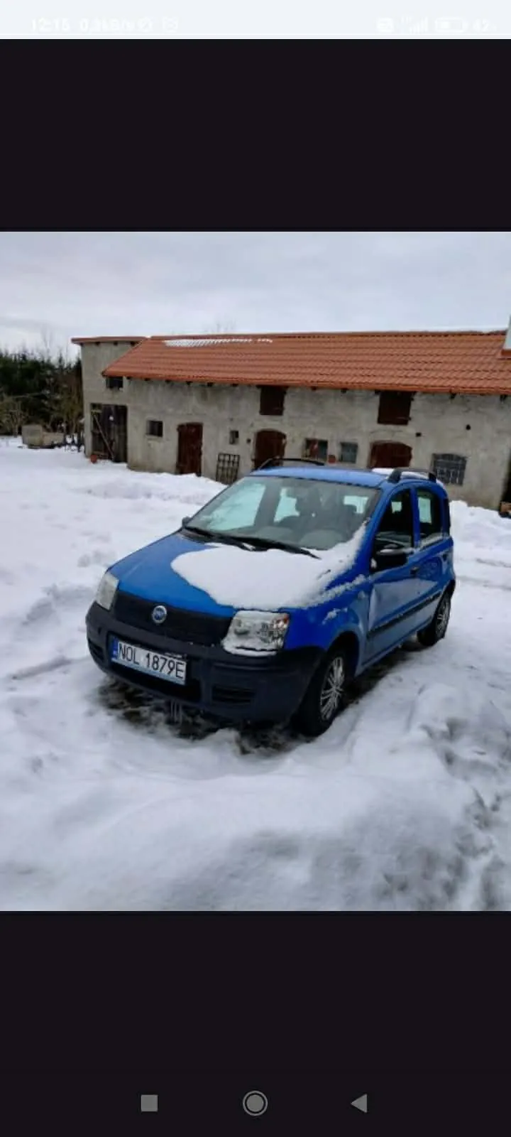 Niebieski samochód z tablicą rejestracyjną NDL 1879E zaparkowany na śnieżnej powierzchni przed budynkiem o białych ścianach i czerwonym dachu. Na dachu budynku oraz na masce samochodu leży śnieg. W tle widoczne są drzwi, okna i częściowo zasłonięte drzewa. Niebo jest zachmurzone.