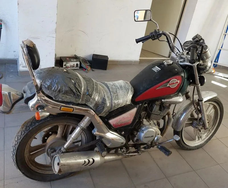 Czarno-czerwony motocykl marki Daewoo z siedzeniem owiniętym przezroczystą folią. Motocykl posiada chromowane elementy, lusterko wsteczne na kierownicy oraz widoczną tablicę z napisem 'DAEWOO' na silniku. W tle biała ściana, płytki podłogowe oraz czarne i srebrne przedmioty, w tym pudełko i urządzenie elektroniczne. Motocykl stoi w pomieszczeniu o jasnym oświetleniu.