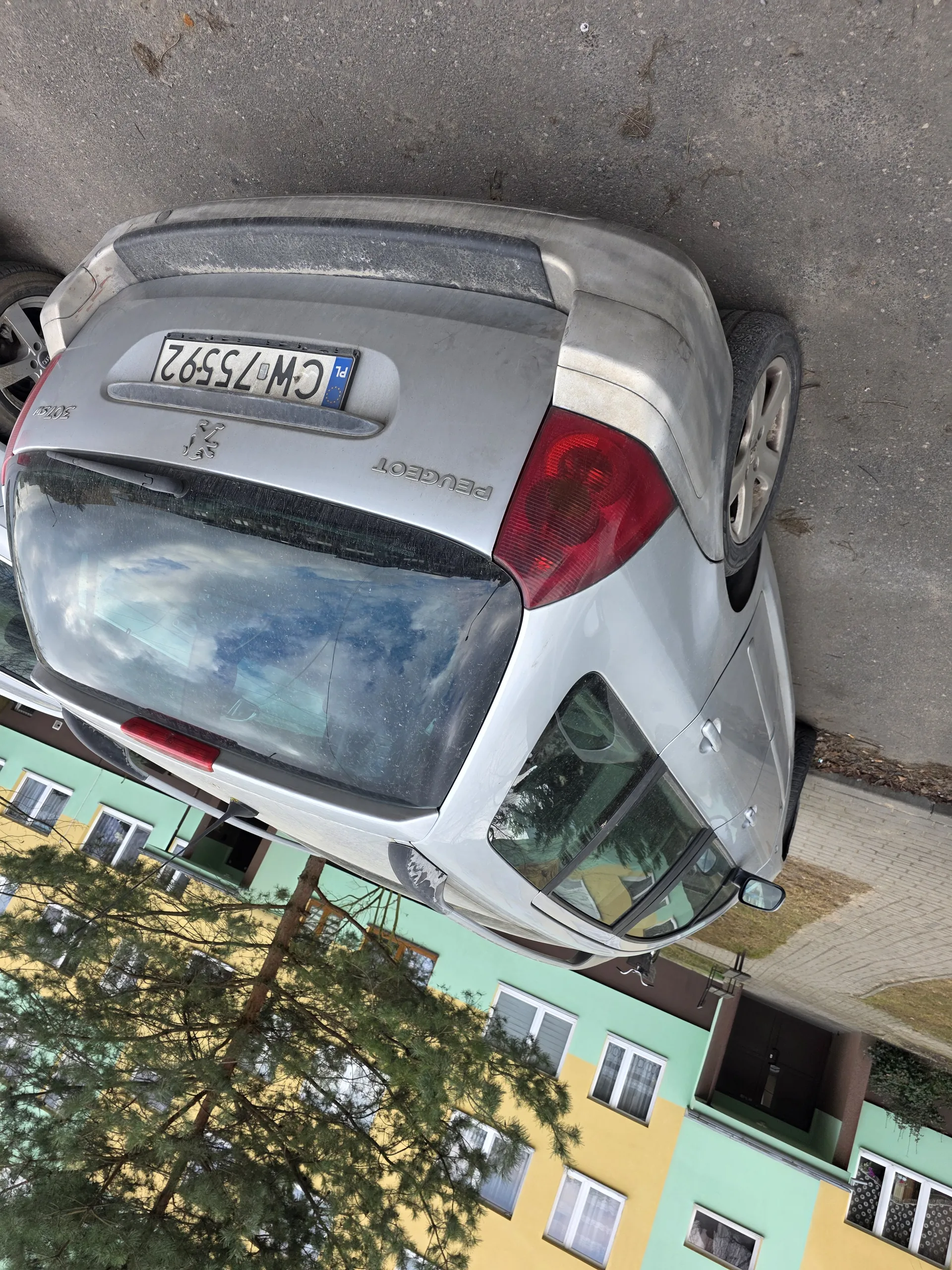 Tylne widok srebrnego samochodu marki Peugeot z tablicą rejestracyjną CW75592 (PL). Samochód zaparkowany na asfaltowej powierzchni. W tle budynki o żółto-zielonych ścianach z oknami oraz drzewo. Widoczne są czerwone światła tylne i logo marki Peugeot.