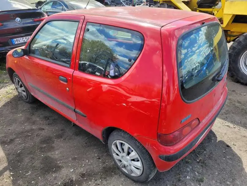 Czerwony samochód Fiat Uno z trzema drzwiami, widoczny z tyłu i boku. Pojazd ma srebrne felgi, lekkie zadrapania na karoserii oraz odbicia nieba i drzew w szybach. W tle: czarny samochód z tablicą rejestracyjną HN C 54, żółta maszyna budowlana oraz inne pojazdy. Podłoga ziemna z fragmentami trawy.