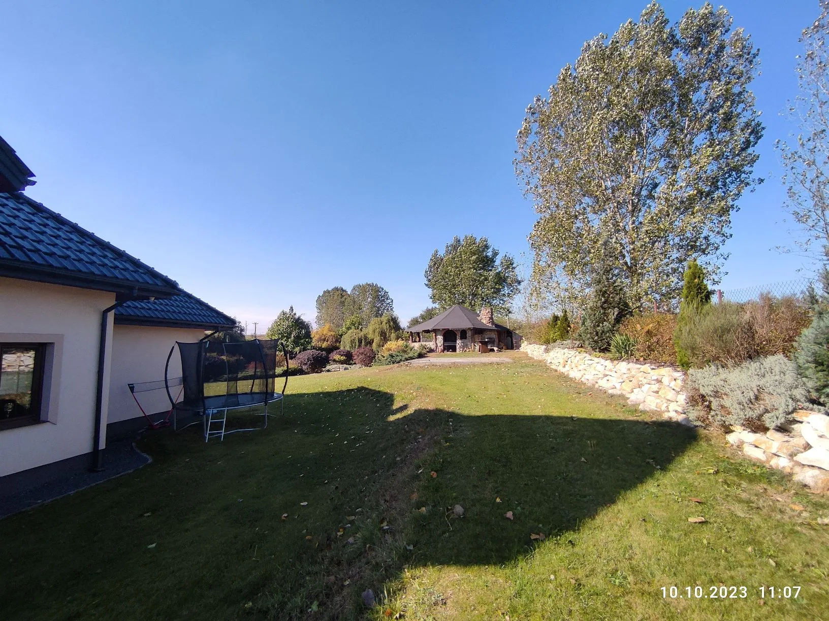 Widok na zadni ogród z zielonym trawnikiem. Po lewej stronie znajduje się fragment domu z ciemnym dachem i jasną elewacją. Na trawniku stoi trampolina z siatką zabezpieczającą. W oddali widoczna jest drewniana altana z dachem, otoczona drzewami i krzewami. Po prawej stronie przebiega niski murek oporowy z jasnych kamieni. W prawym dolnym rogu znajduje się data i godzina: 10.10.2023 11:07.