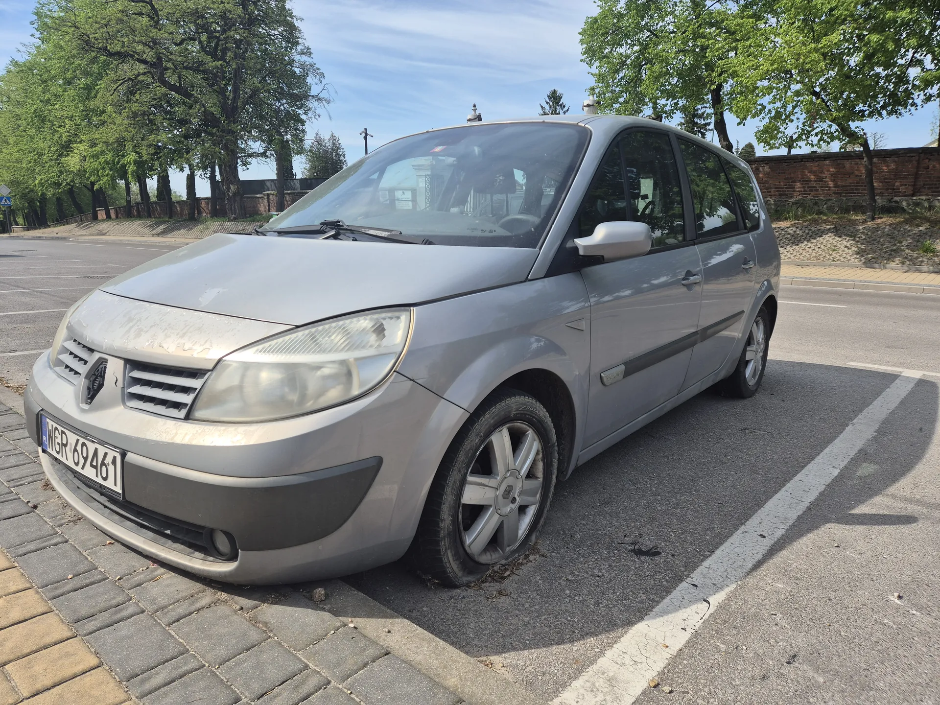 Srebrny samochód osobowy marki Renault zaparkowany na poboczu drogi. Widoczna tablica rejestracyjna z numerem WGR 69461. W tle drzewa, ceglany mur, chodnik z kostki brukowej oraz droga z białymi liniami. Niebo jest częściowo zachmurzone.