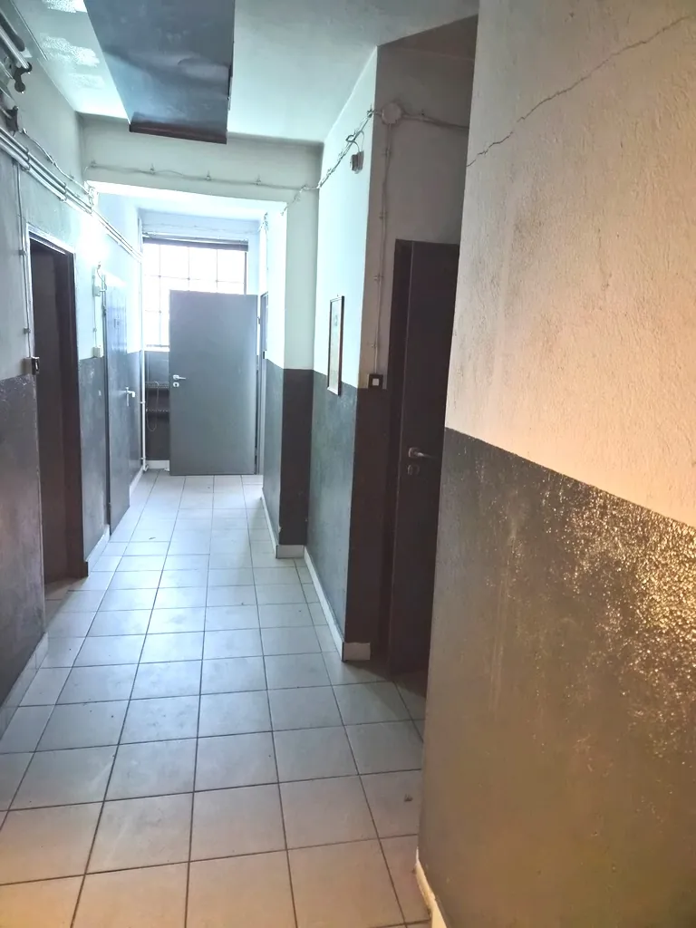 Zdjęcie przedstawia korytarz w budynku z jasnymi ścianami i podłogą wyłożoną szarymi płytkami. Wzdłuż korytarza znajdują się ciemne, prawdopodobnie drewniane lub metalowe elementy konstrukcyjne oraz drzwi. Na suficie widoczne jest oświetlenie liniowe. Obraz jest zorientowany pionowo, mimo że przedstawia przestrzeń poziomą.