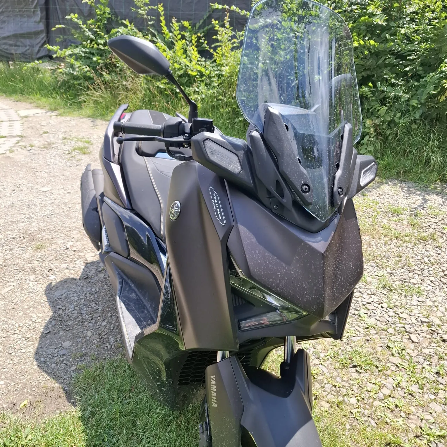 Czarny skuter marki Yamaha zaparkowany na piaszczystej drodze obok zielonej roślinności. Pojazd posiada wysoką, przezroczystą szybę ochronną, lusterka boczne i widoczne logo Yamaha na przedniej owiewce oraz błotniku.