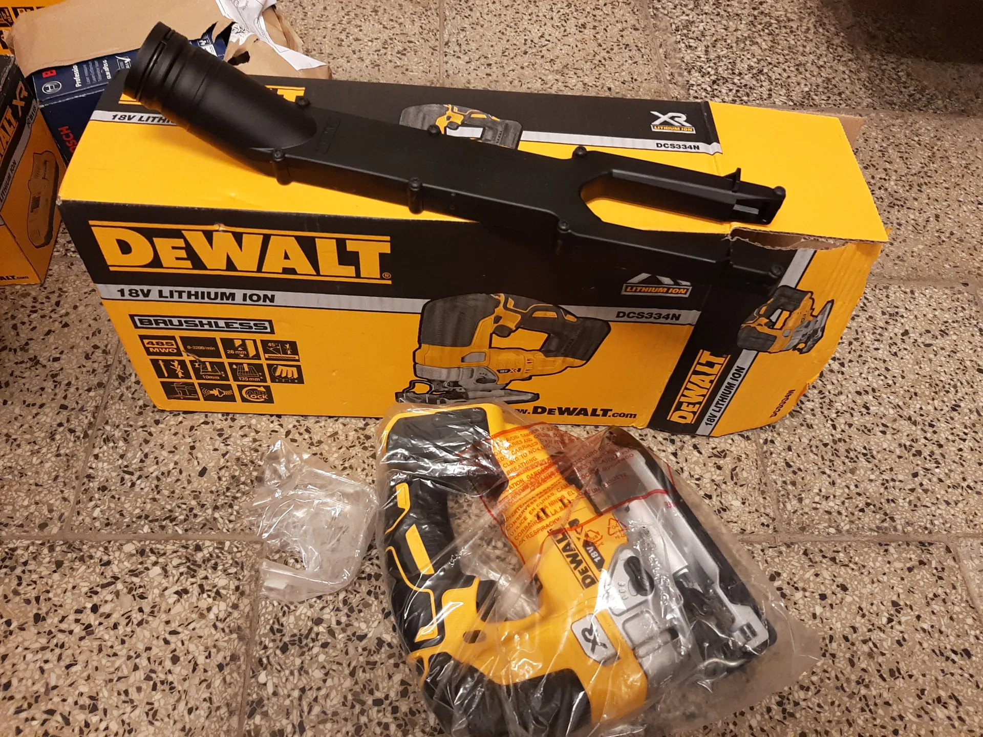 Zdjęcie przedstawia żółto-czarną pudełko marki DEWALT z napisem 18V LITHIUM ION, BRUSHLESS oraz oznaczeniem modelu DCS334N. Na pudełku widoczne są ikony specyfikacji technicznych i ilustracja narzędzia. Na pudełku leży czarna rękojeść lub akcesorium. Przed pudełkiem znajduje się opakowane w przezroczystą folię żółto-czarne narzędzie z widocznym logo DEWALT i napisem XR, obok mały woreczek z plastiku. Tło stanowi podłoga z kafelkami w kolorze beżowym z czarnymi kropkami. Na pudełku widoczny jest również adres www.DEWALT.com.