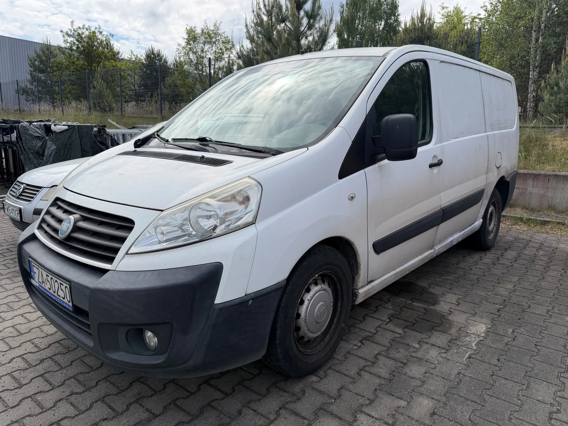 Biały furgon marki Fiat zaparkowany na brukowanej powierzchni. Widoczna tablica rejestracyjna FZA 50250. W tle drugi pojazd częściowo zakryty, ogrodzenie, drzewa i przemysłowe budynki. Niebo częściowo zachmurzone. Furgon ma czarne elementy na zderzaku i boku, a przednie szyby odbijają chmury.