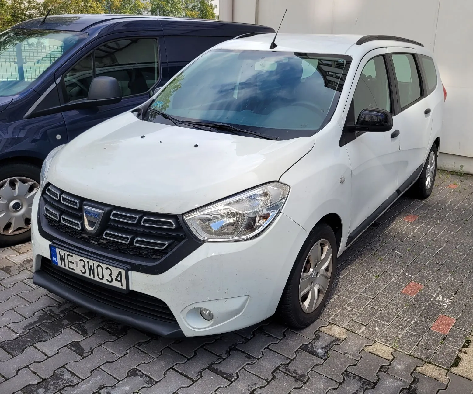 Biały samochód marki Dacia zaparkowany na kostce brukowej obok ciemnoniebieskiego vana. Widoczna jest przednia część i prawy bok białego auta z tablicą rejestracyjną o numerze WE 3WO34.