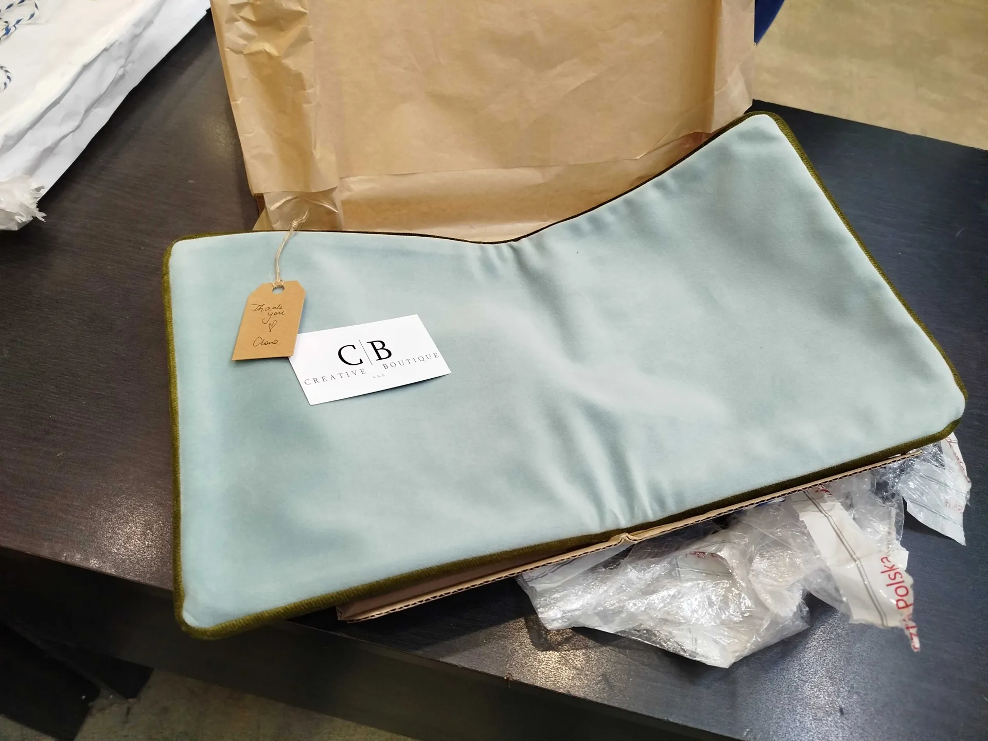 Na ciemnym stole leży jasnoniebieska tkanina z zielonym wykończeniem, na której umieszczono białą kartkę z napisem 'C|B CREATIVE BOUTIQUE' oraz brązowy etykietę z napisem 'Thank you' i sercem. Za tkaniną znajduje się brązowa papierowa torba. W dolnej części obrazu widoczne są przezroczyste opakowania z plastiku oraz fragment białego papieru z czerwonym napisem 'Polska'.