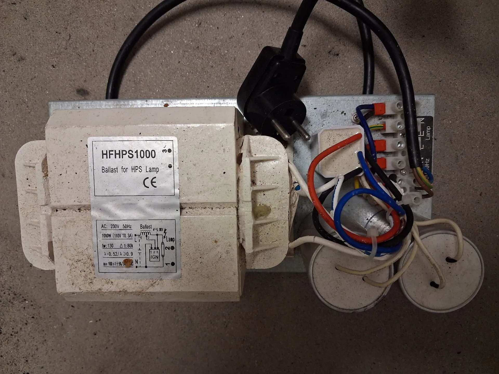Urządzenie elektryczne zamontowane na metalowej podstawie. Po lewej stronie znajduje się biały, prostokątny dławik z etykietą zawierającą tekst: 'HFHPS1000 Ballast for HPS Lamp CE' oraz schemat techniczny z danymi: 'AC 230V 50Hz', '1000W (10kV 10.3A)'. Po prawej stronie widoczne są kolorowe przewody elektryczne (niebieski, brązowy, pomarańczowy, czarny) podłączone do kostki zaciskowej oraz dwa białe kondensatory cylindryczne. Do urządzenia podłączony jest czarny kabel z wtyczką sieciową typu C.