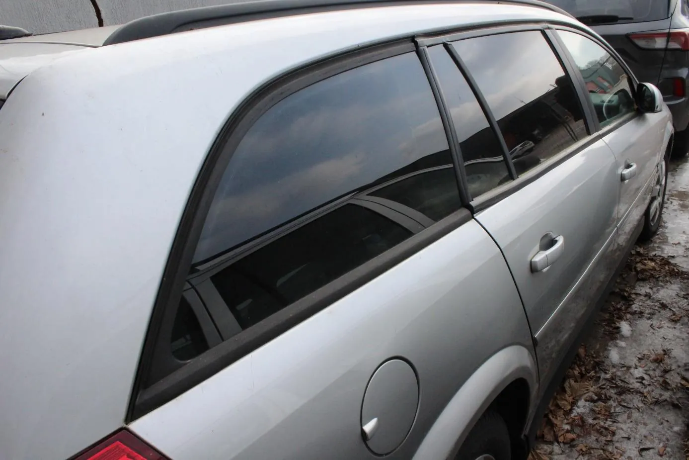 Zbliżenie na srebrny samochód typu kombi, widok z tyłu i boku. Widoczne są przyciemniane szyby, klamki drzwi, pokrywa zbiornika paliwa oraz czerwone światło tylnego. Samochód zaparkowany na nawierzchni z suchymi liśćmi i resztkami śniegu. W tle częściowo widoczny inny, ciemny samochód.