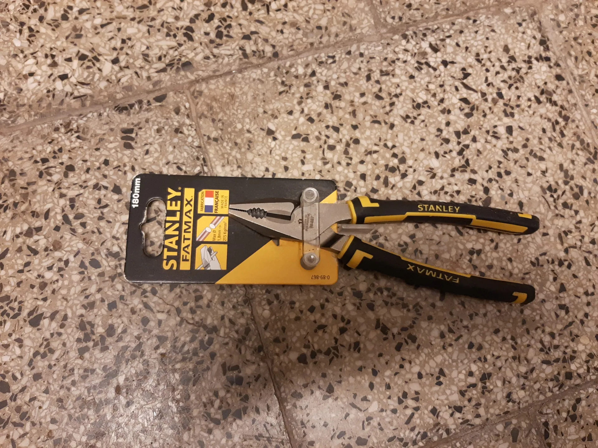 Otwarte szczypce Stanley FatMax o długości 180 mm w opakowaniu. Opakowanie czarno-żółte z napisem „STANLEY FATMAX” oraz informacją „180mm” po lewej stronie. Na opakowaniu widoczne są małe ikony, flaga Francji i napis „FRANÇAISE”. Szczypce mają czarno-żółte uchwyty z napisami „STANLEY” i „FATMAX”. Tło stanowi posadzka z kruszywem w kolorach beżowym i czarnym.