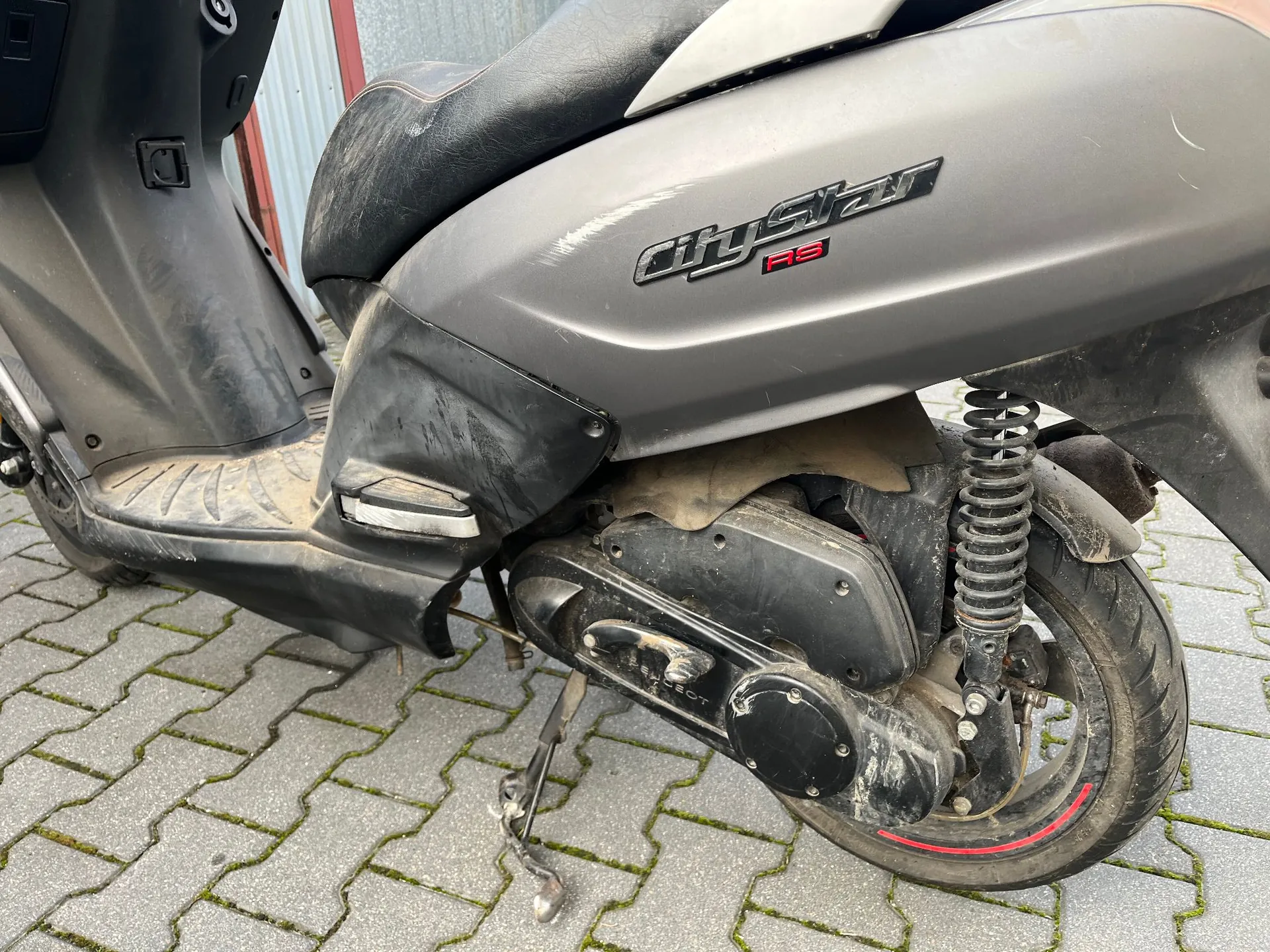 Zbliżenie na prawy bok szarego skutera zaparkowanego na kostce brukowej. Widoczny jest fragment siedzenia, nadwozie z napisem „CityStar RS”, amortyzator oraz tylne koło. Pojazd jest zakurzony i posiada widoczne zarysowania na plastikowych elementach.