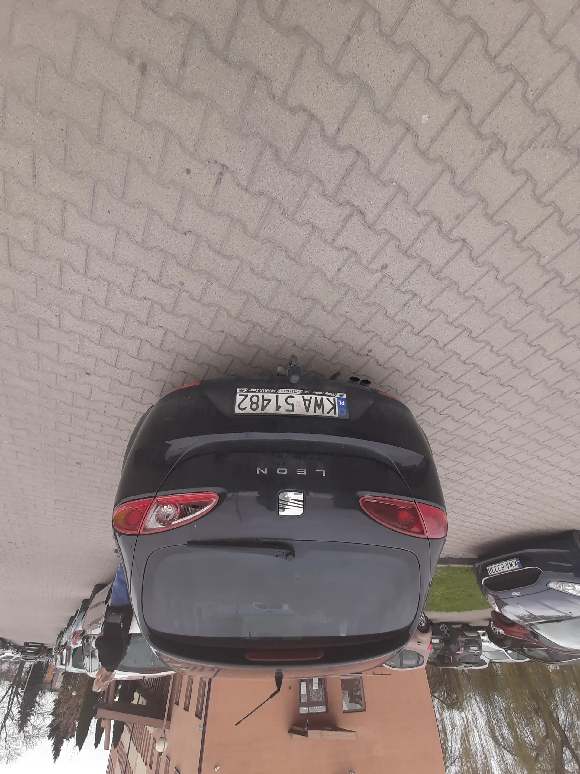 Czarny samochód SEAT Leon zaparkowany na brukowanej powierzchni. Widoczna tablica rejestracyjna KWA 51482. W tle inne zaparkowane samochody, pomarańczowy budynek, drzewa oraz osoba idąca. Droga wybrukowana, po lewej stronie zieleń.