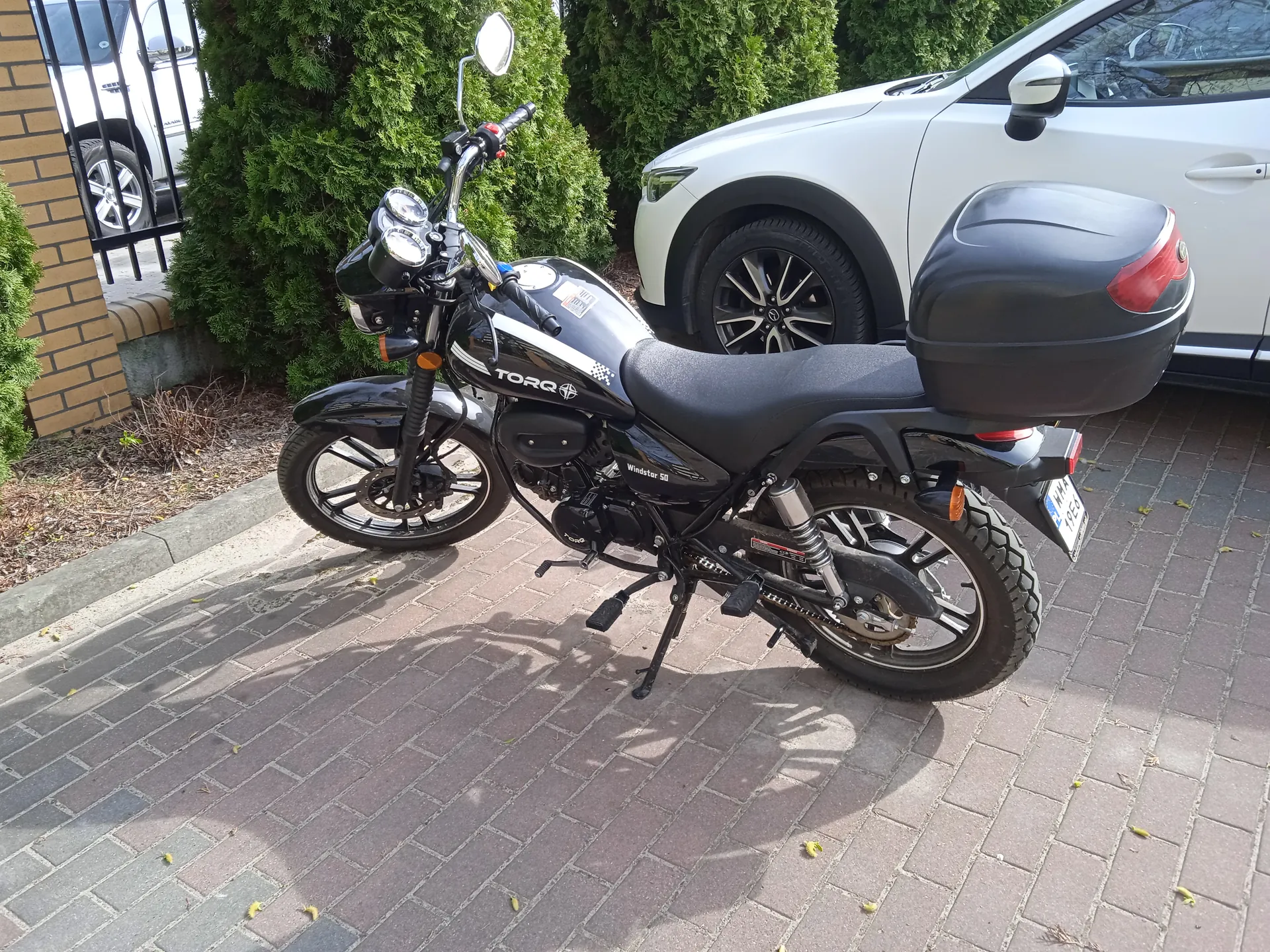 Czarny motocykl marki TORQ zaparkowany na brukowanej powierzchni. Motocykl posiada czarny bagażnik, tablicę rejestracyjną oraz widoczny napis TORQ na baku. W tle biały samochód osobowy, zielone krzewy, ceglana ściana i metalowa brama. Na ziemi leżą rozrzucone żółte liście.