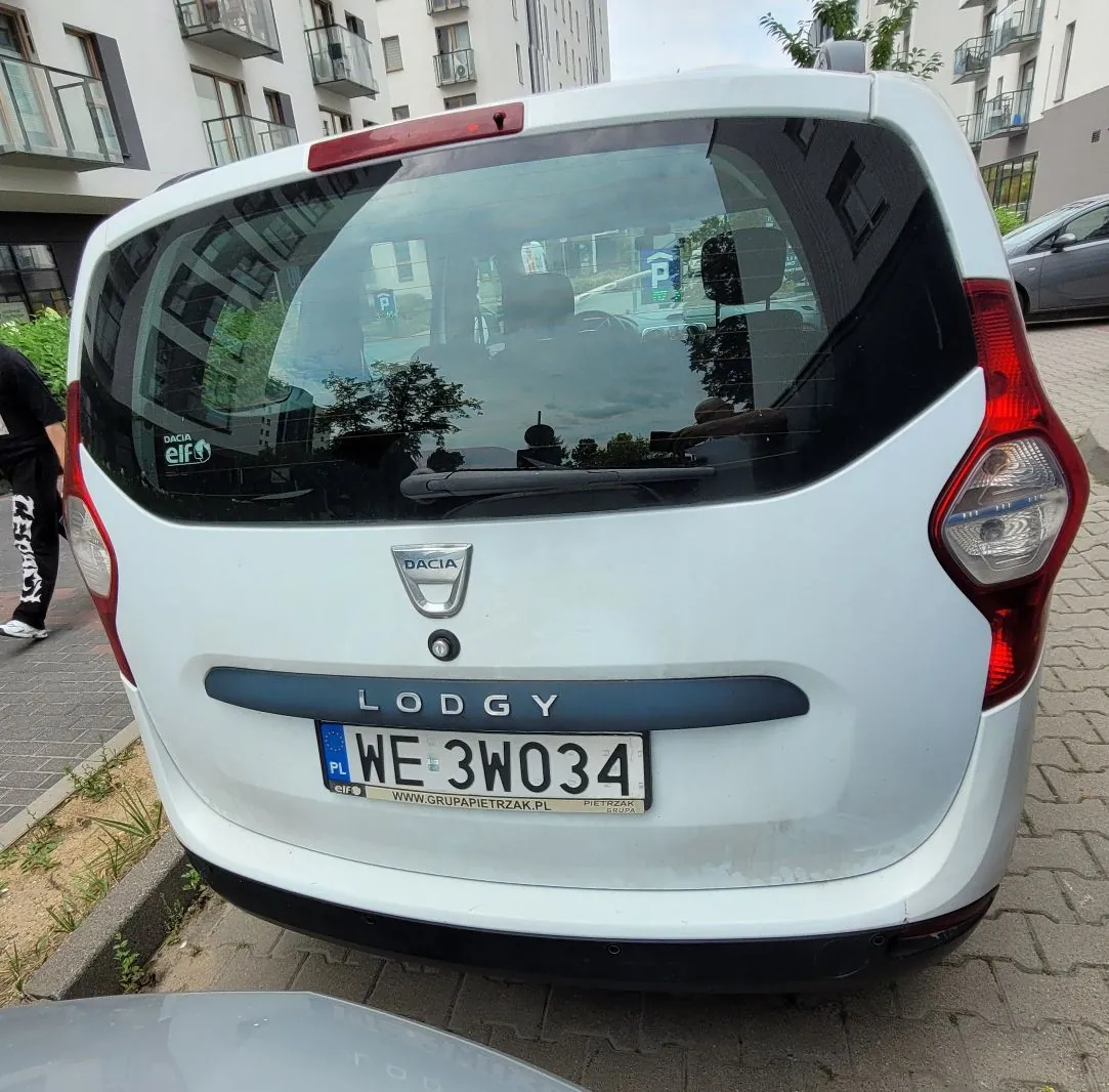 Tylny widok białego samochodu marki Dacia, model Lodgy. Na klapie bagażnika znajduje się logo Dacia oraz napis LODGY. Samochód ma polską tablicę rejestracyjną o numerze WE 3W034. Na szybie tylnej widoczna jest naklejka 'elf'. W tle widać nowoczesne, jasne budynki mieszkalne z balkonami.