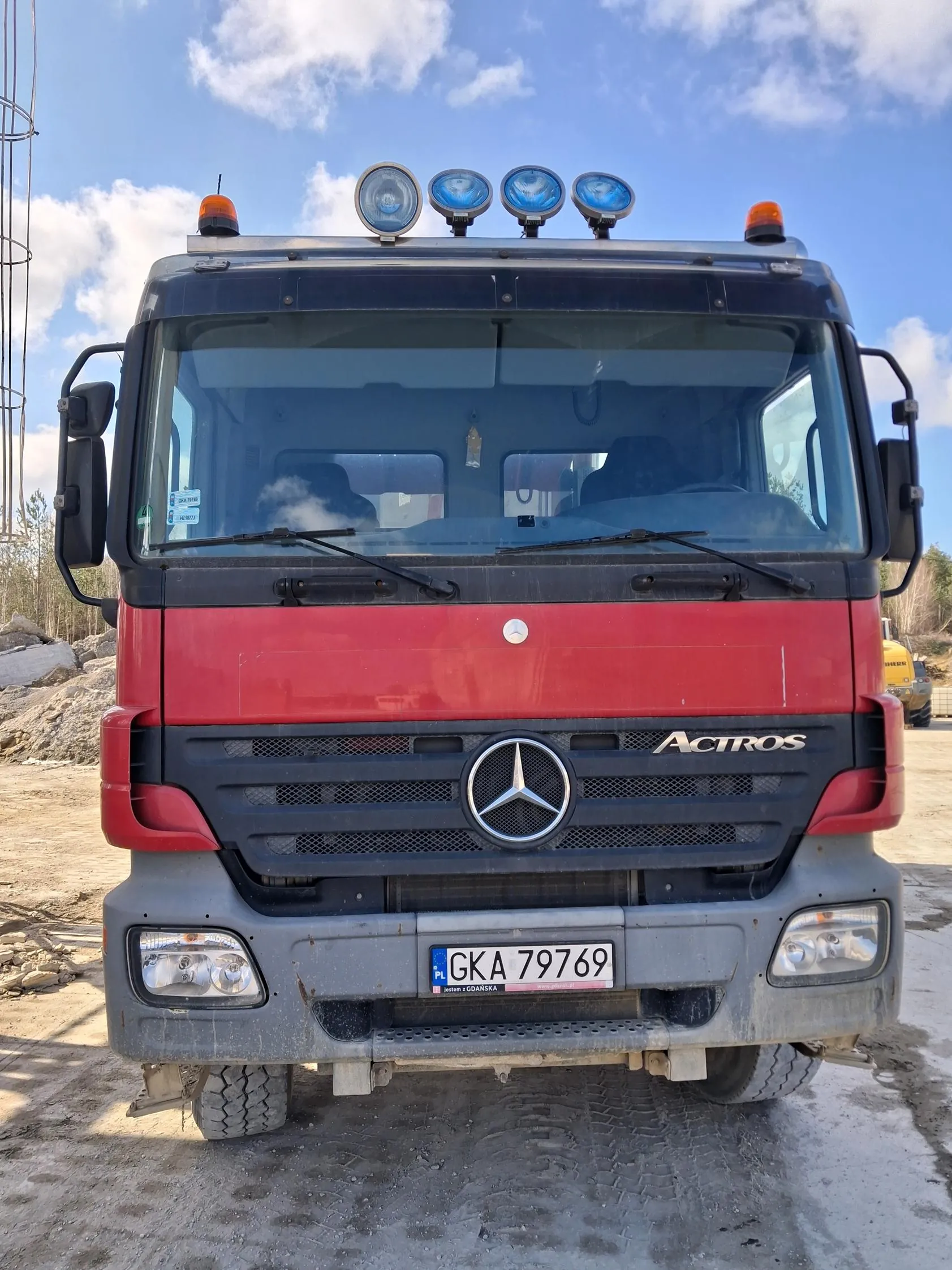 Przednia część czerwono-czarnego ciężarówki Mercedes-Benz Actros z widocznym logo trójgwiazdy na chłodnicy. Na masce umieszczono cztery niebieskie reflektory oraz dwa pomarańczowe sygnalizatory. Tablica rejestracyjna PL GKA 79769 z napisem "Jestem z GDAŃSKA" na dole. Tło przedstawia teren budowlany z kamieniami, żółtym koparką i niebem z chmurami. Widoczne są również lusterka boczne, przednie szyby z wycieraczkami oraz fragmenty siedzeń w kabinie.