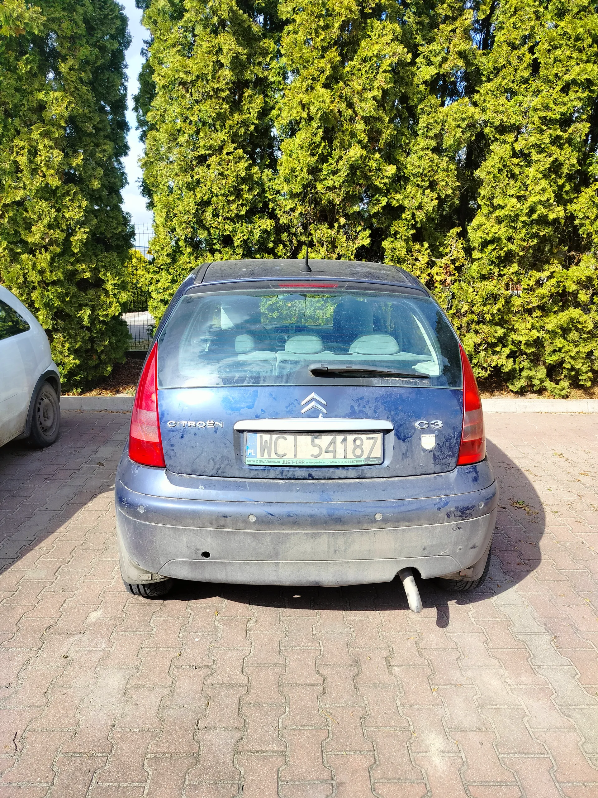 Tylne widok niebieskiego samochodu Citroën C3 z brudną karoserią zaparkowanego na brukowanej powierzchni. Widoczna tablica rejestracyjna PL WCI 54187. Po lewej stronie częściowo widoczny biały samochód. Tło stanowią wysokie zielone drzewa i jasne niebo. Samochód ma czerwone światła tylne, wycieraczkę na tylnej szybie oraz widoczny wydech.