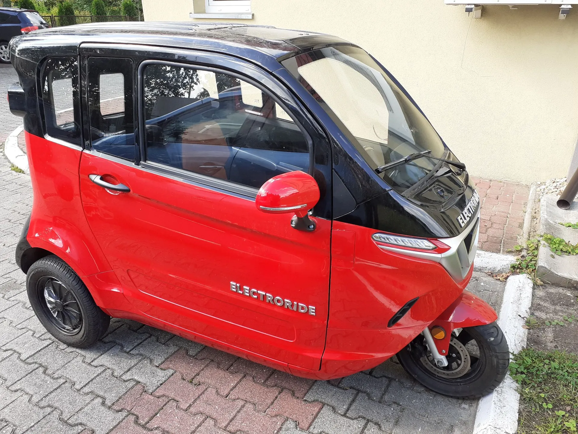 Mały, dwukolorowy pojazd elektryczny zaparkowany na kostce brukowej. Karoseria pojazdu jest czerwona w dolnej części i czarna w górnej części, w tym dach i słupki. Na boku pojazdu widnieje biały napis „ELECTRORIDE”. Pojazd ma cztery małe koła z czarnymi felgami i czerwone lusterko boczne.