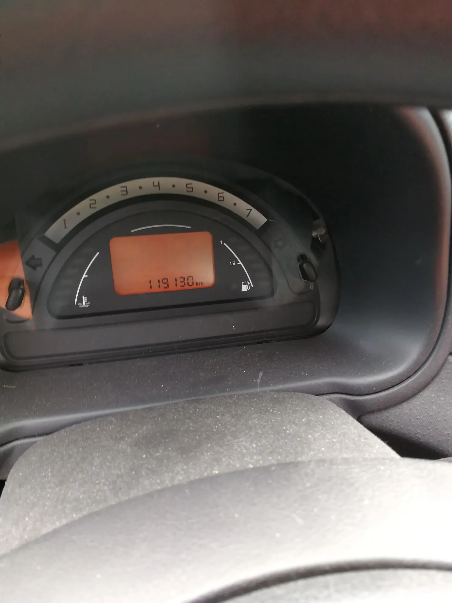 Zbliżenie na deskę rozdzielczą samochodu. Widoczny jest licznik kilometrów z pomarańczowym wyświetlaczem cyfrowym wskazującym wartość 119 130 km. Nad wyświetlaczem znajduje się skala z numerami od 1 do 6. Po prawej stronie widoczny jest wskaźnik poziomu paliwa z ikoną dystrybutora.