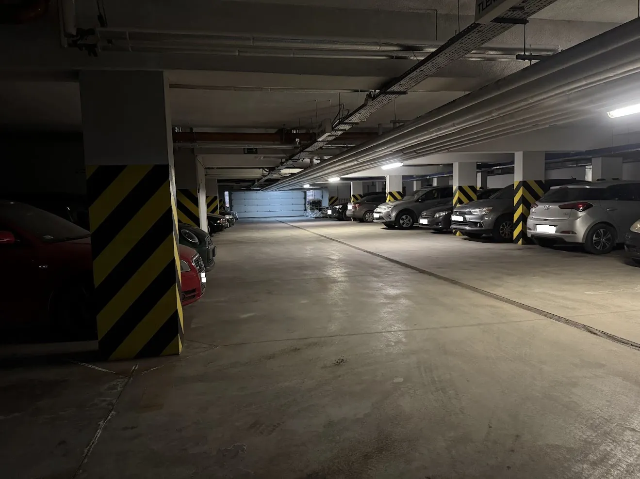 Podziemny parking z rzędami zaparkowanych samochodów. Po lewej stronie widoczny jest fragment czerwonego auta oraz betonowy filar z żółto-czarnymi pasami ostrzegawczymi. Po prawej stronie zaparkowane są srebrne i białe samochody osobowe. Sufit jest niski, z widocznymi rurami instalacyjnymi i liniowymi lampami oświetlającymi betonową podłogę.