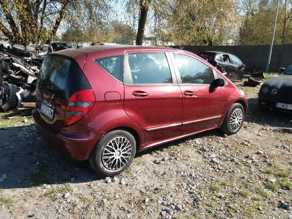 Czerwony samochód osobowy Mercedes-Benz B-Klasa zaparkowany na żwirowym terenie. Widoczna tablica rejestracyjna HJ 171 AM. W tle zniszczone pojazdy, w tym stos wraków po lewej stronie i czarny samochód z tablicą DW 5Y po prawej. Tło stanowią drzewa, betonowy mur i jasne niebo. Podłoże pokryte kamieniami i rzadką trawą.