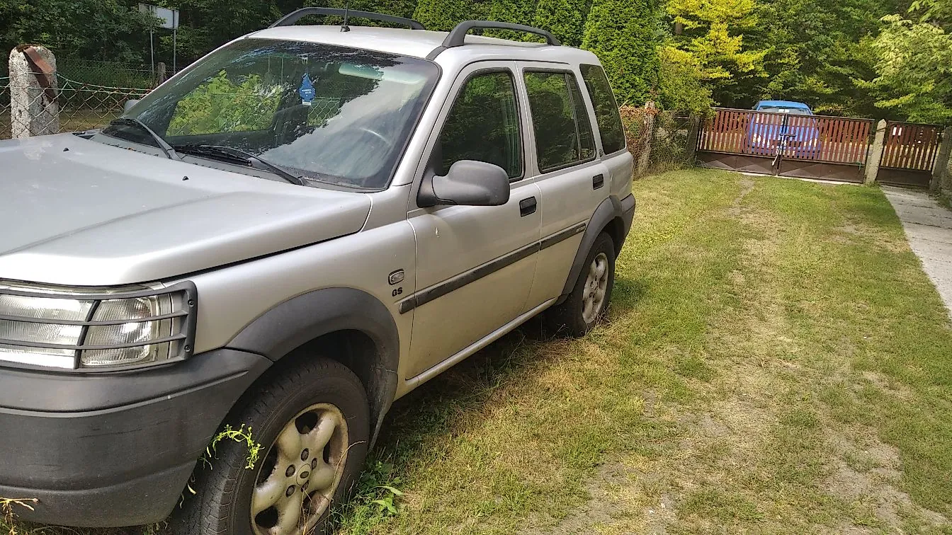 Srebrny samochód typu kombi zaparkowany na trawiastym podjeździe. Pojazd jest widoczny z przodu i z boku, posiada czarne plastikowe osłony nadkoli i progi oraz relingi dachowe. W tle widać zielone drzewa, krzewy oraz drewniany płot, za którym stoi niebieski samochód.