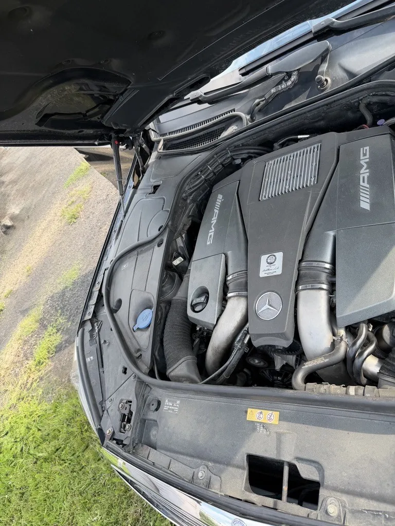 Widok z góry na komorę silnika samochodu marki Mercedes-AMG. Silnik posiada czarną osłonę z napisem 'AMG' po obu stronach oraz logo Mercedesa na środku. Widoczne są rury dolotowe, przewody i elementy konstrukcyjne samochodu. Maska samochodu jest otwarta i widoczna w górnej części kadru.