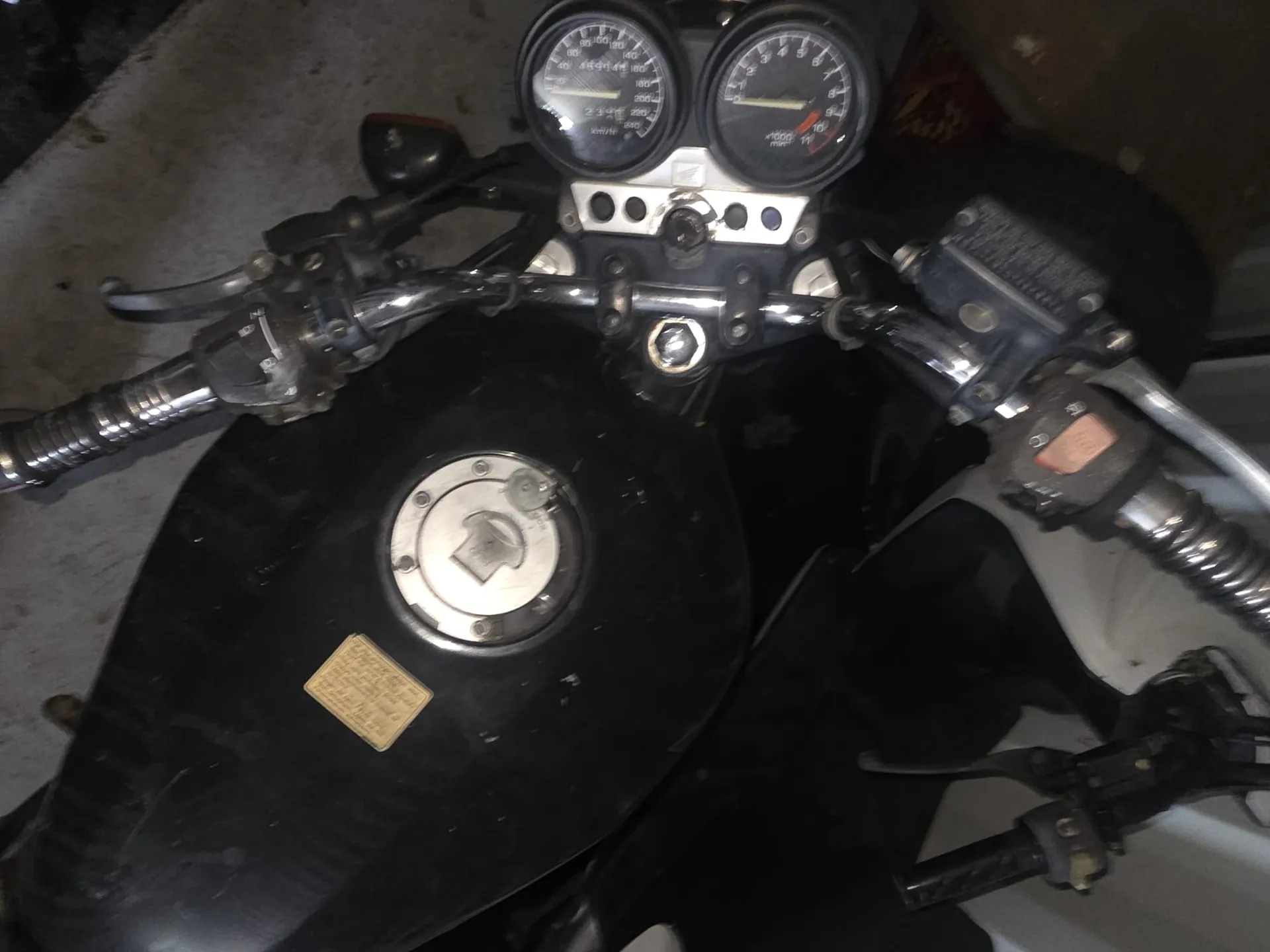 Zbliżenie na kierownicę motocykla z czarnym zbiornikiem paliwa. Widoczne są dwa wskaźniki: górny z czerwoną strefą i dolny z zakresem do 160. Na zbiorniku znajduje się srebrna pokrywka i żółty etykietka z nieczytelnym tekstem. Kierownica wyposażona w czarne dźwignie, przyciski i metalowe elementy. Tło stanowi szara podłoga z widocznymi plamami.
