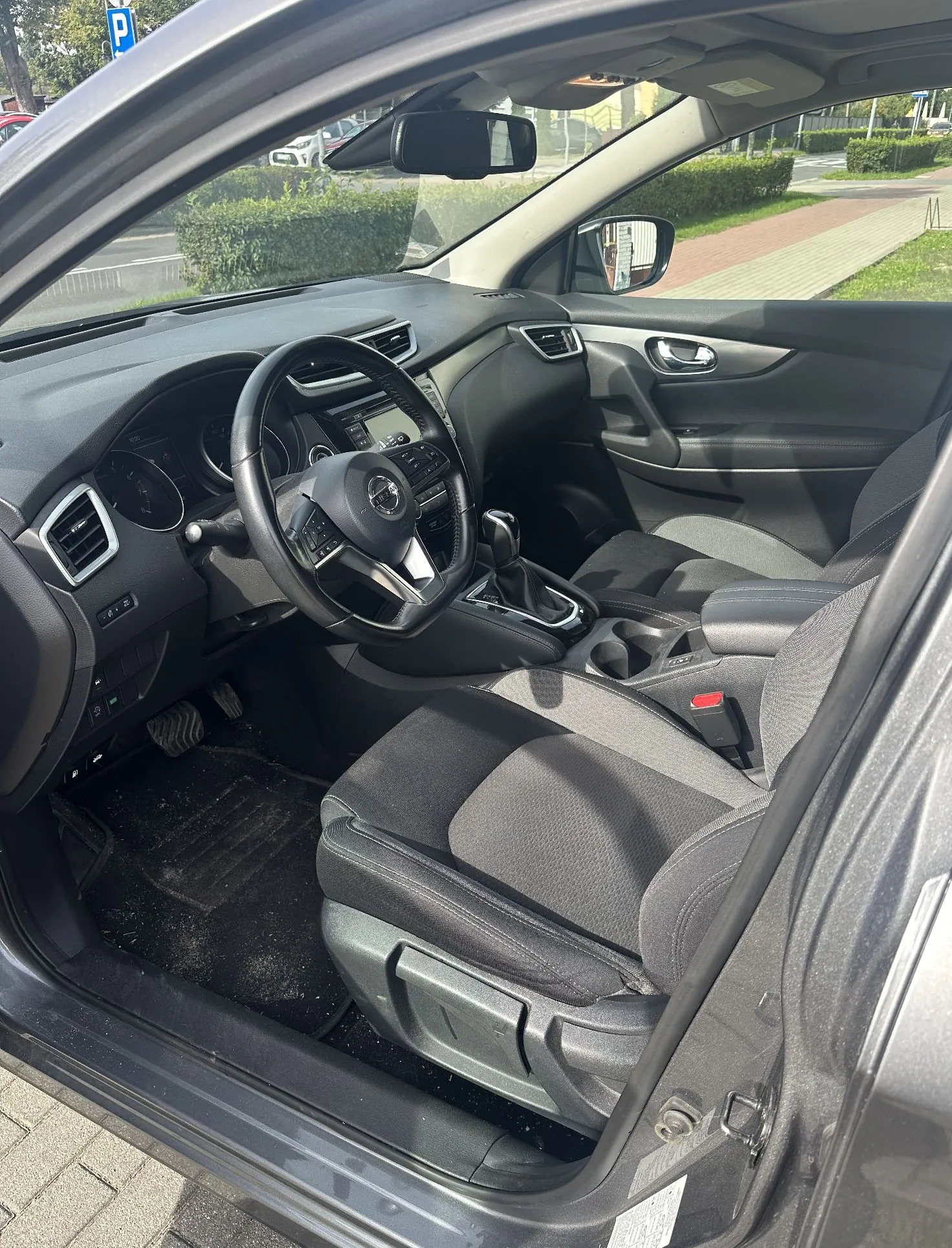Wnętrze samochodu widziane z perspektywy otwartych przednich drzwi kierowcy. Widoczna jest czarna tapicerka materiałowa na fotelach, czarna kierownica z logo marki Opel w centrum, deska rozdzielcza z zegarami i systemem audio oraz lewarek zmiany biegów. Przez przednią szybę i lusterka widać fragmenty ulicy, chodnika i zieleni.