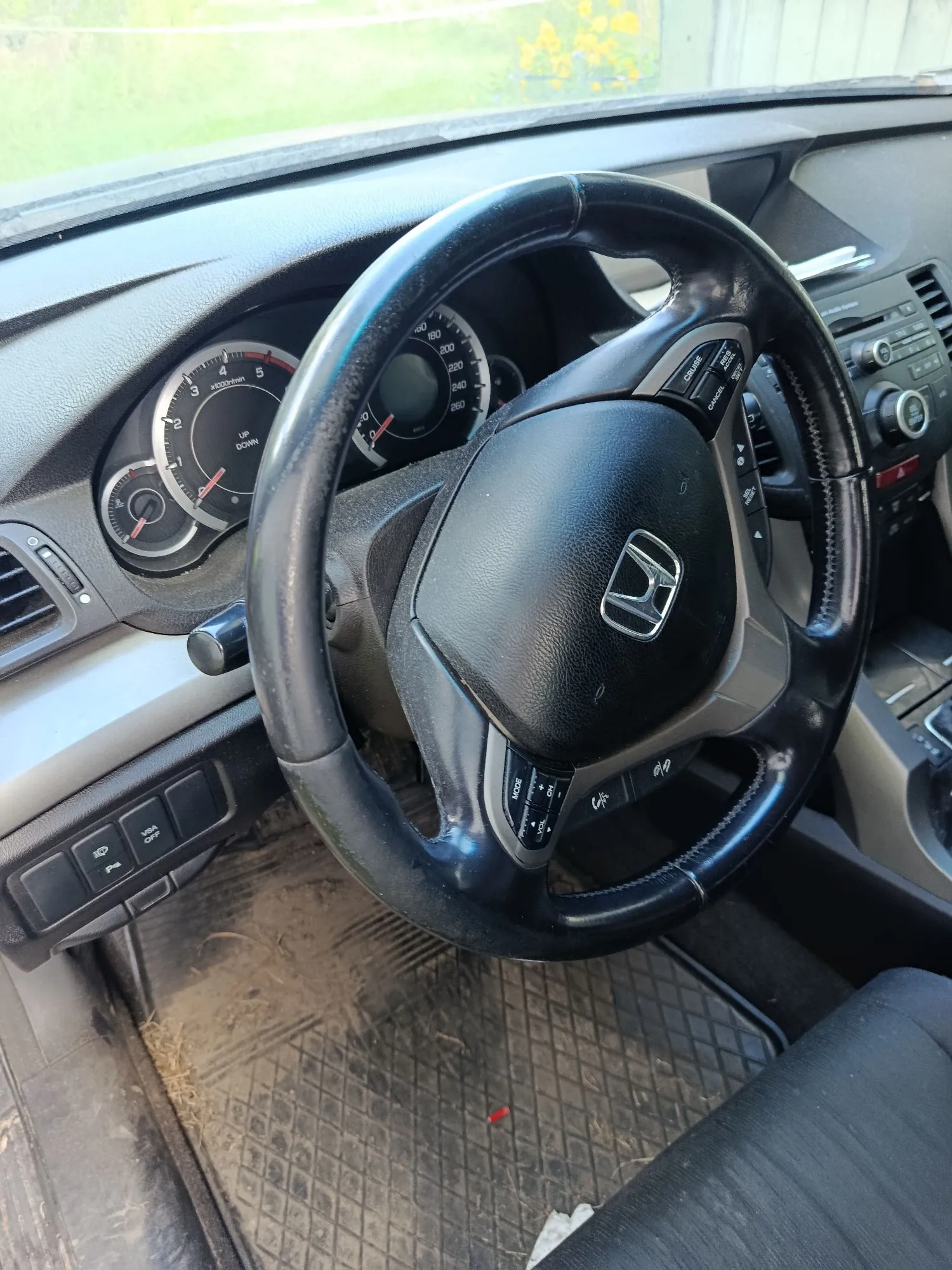 Wnętrze samochodu z perspektywy kierowcy. Widoczna jest czarna kierownica z logo marki Honda na środku, deska rozdzielcza z analogowymi wskaźnikami prędkości i obrotomierzem oraz fragmenty konsoli środkowej. Na podłodze znajduje się gumowa mata z widocznymi zabrudzeniami w postaci piasku i suchych liści.
