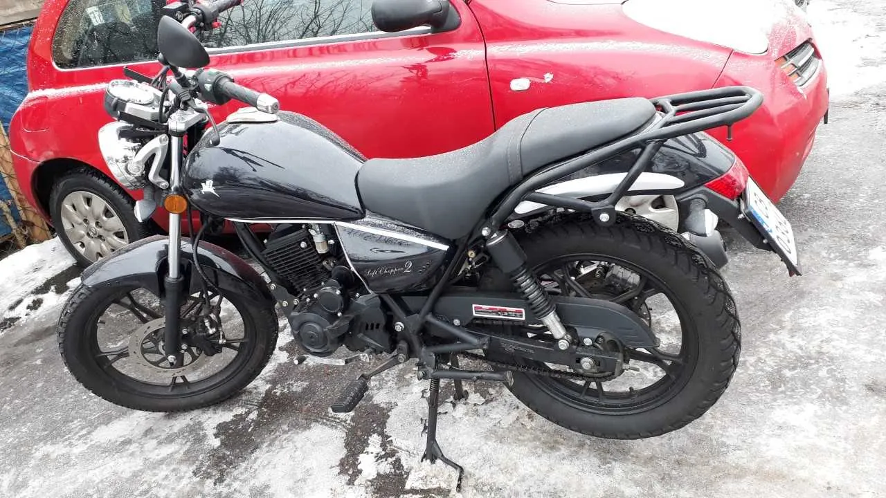 Czarny motocykl z widocznym napisem "Left Chopper 2" na boku, czarnym siedzeniem i tablicą rejestracyjną, zaparkowany na śnieżnej nawierzchni. Za nim czerwony samochód osobowy. Na tle drzewa i niebieska konstrukcja. Śnieg na ziemi i częściach pojazdów.