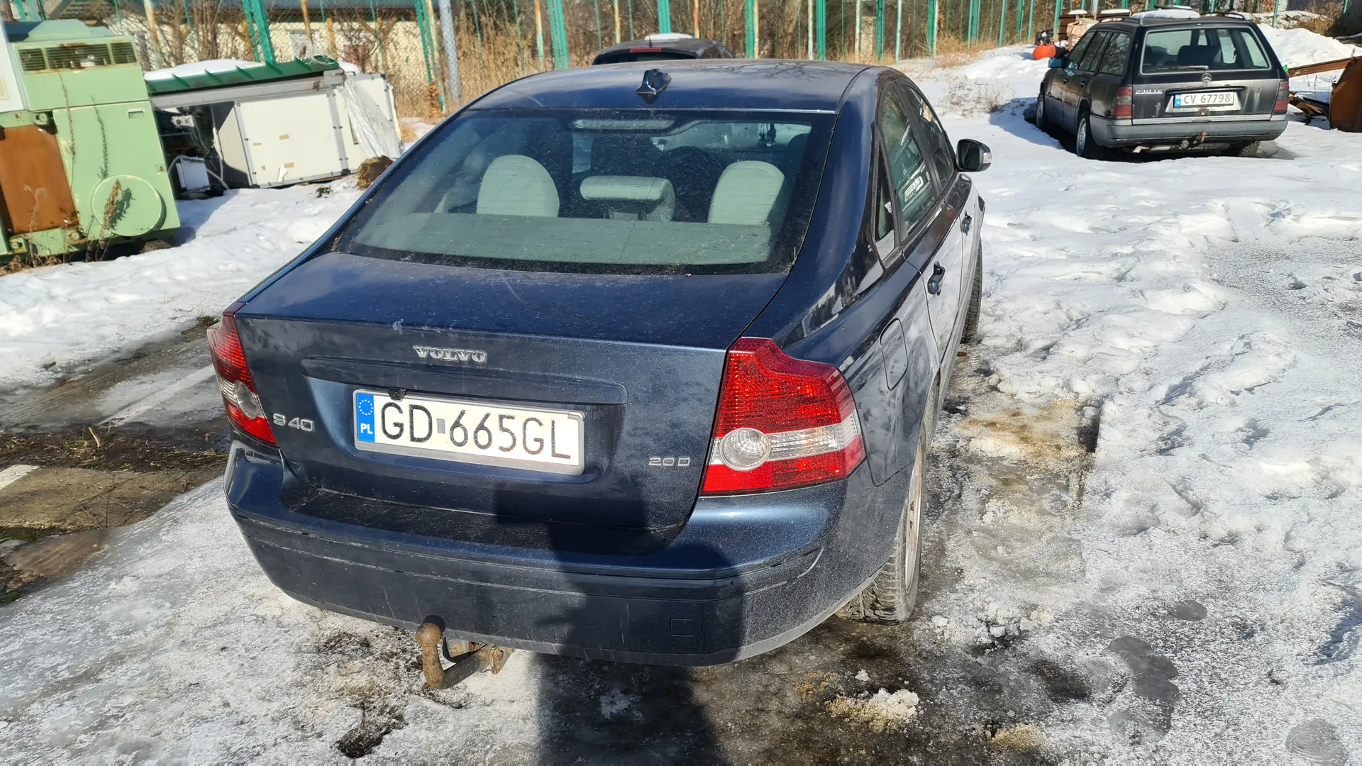 Z tyłu widoczny ciemnoniebieski samochód Volvo S40 z tablicą rejestracyjną PL GD-665GL. Na tle śnieżnego terenu stoją inne pojazdy, w tym szary samochód z tablicą CV 67798. W tle zielone ogrodzenie, przemysłowe urządzenia i suche krzewy. Samochód ma oznaczenia S40 i 200 na bagażniku, a podwozie częściowo pokryte śniegiem i lodem.