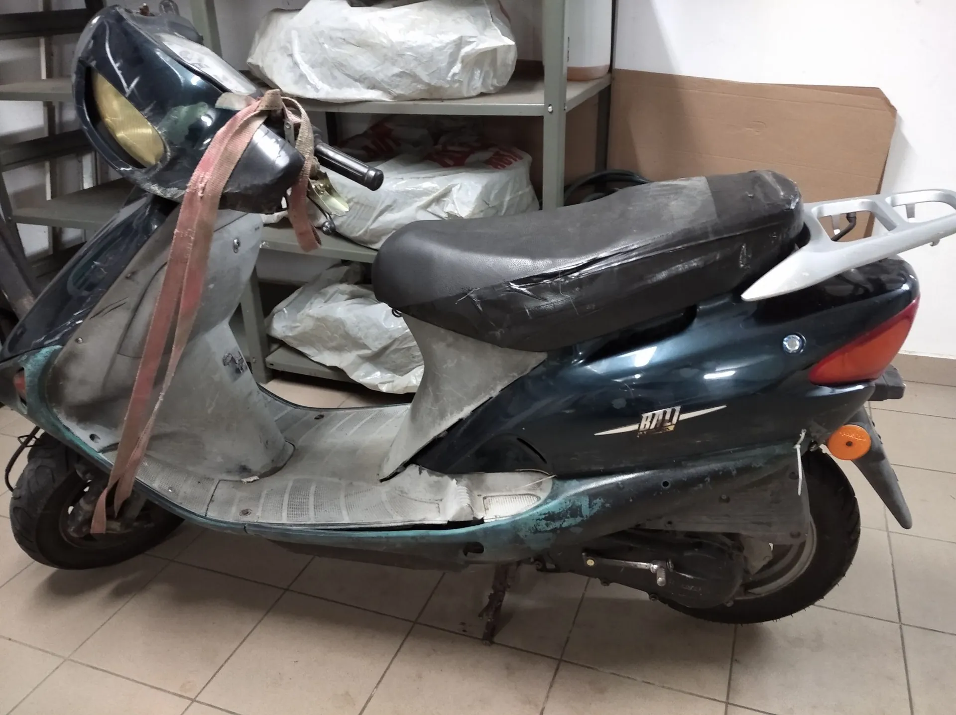 Ciemnoniebieski skuter zaparkowany w pomieszczeniu na kafelkach. Pojazd jest w złym stanie technicznym, z widocznymi uszkodzeniami i zabrudzeniami. Przez kierownicę i przód skutera przechodzi czerwony pas transportowy. W tle widoczny jest metalowy regał z białymi workami oraz kawałek brązowego kartonu.