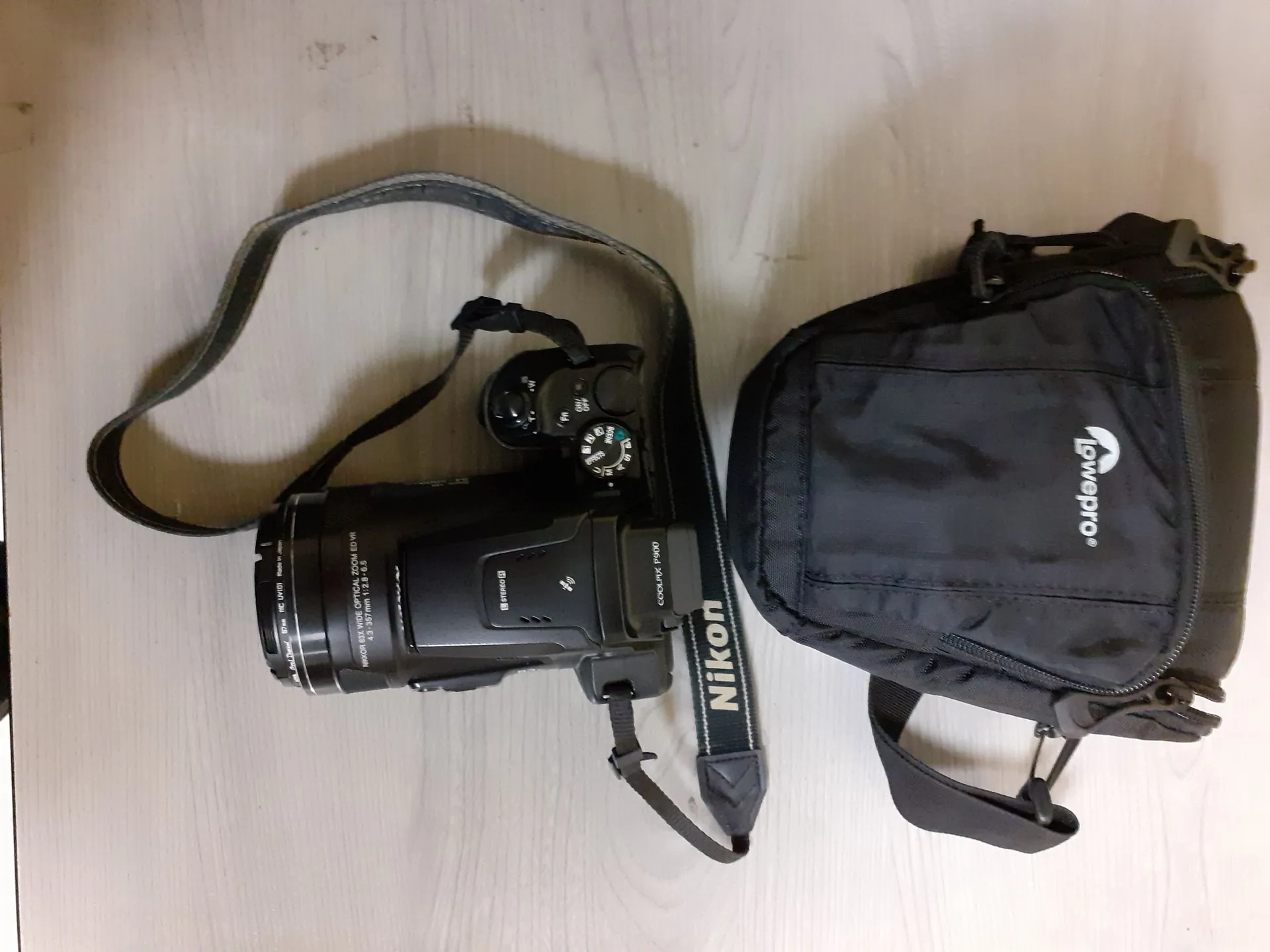 Czarny aparat fotograficzny marki Nikon z obiektywem, leżący na jasnej, drewnianej powierzchni obok czarnej torby fotograficznej marki Lowepro. Aparat ma przymocowany czarny pasek na ramię. Na obiektywie widoczny jest napis 'NIKKOR ZOOM DIGITAL OPTICAL'.