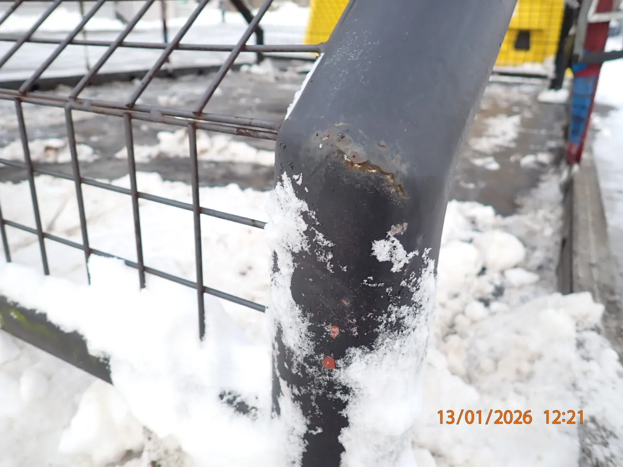 Czarna metalowa rura z widocznymi śladami korozji i śniegiem na powierzchni. W tle metalowa siatka ogrodzeniowa, śnieg na ziemi oraz żółta konstrukcja. W prawym dolnym rogu widoczny pomarańczowy znaczek z datą i czasem: 13/01/2026 12:21.