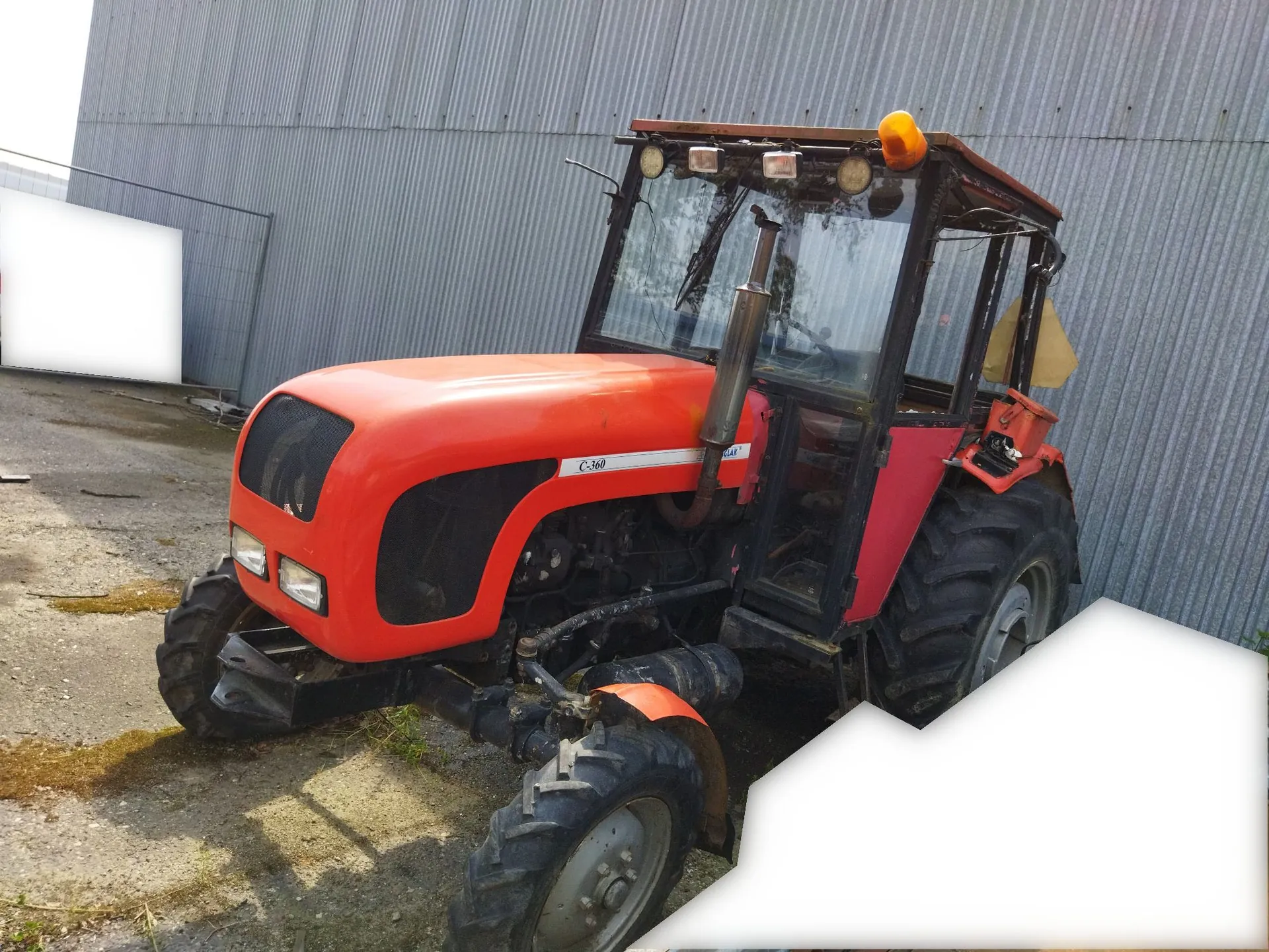 Pomarańczowy traktor marki Ursus, model C-360, zaparkowany na betonowym placu przed budynkiem z szarej, falistej blachy. Traktor posiada czarną kabinę z szybami oraz duże, gumowe koła z charakterystycznym bieżnikiem.
