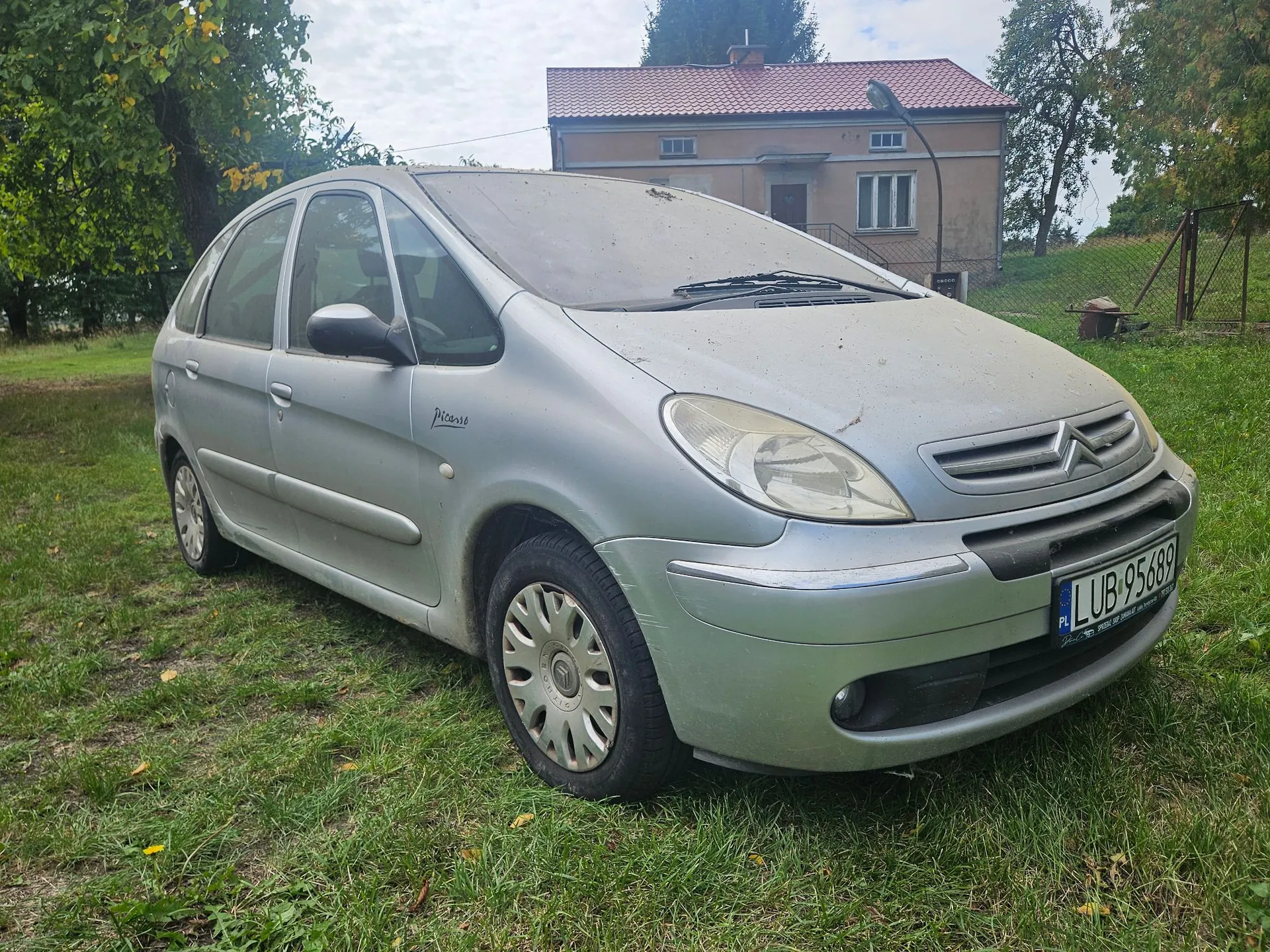 Srebrny samochód osobowy marki Citroën Picasso zaparkowany na trawiastym terenie. Pojazd jest zakurzony i posiada polską tablicę rejestracyjną o numerze LUB 95689. W tle widoczny jest jednopiętrowy dom z czerwonym dachem oraz zielone drzewa.