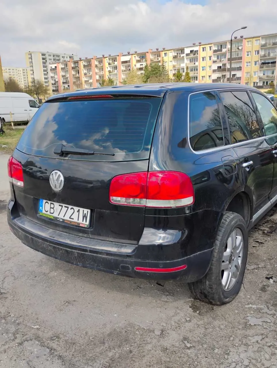 Czarny samochód osobowy typu SUV marki Volkswagen widziany od tyłu. Na tylnej szybie refleksje chmur, czerwone światła tylnego oświetlenia i tablica rejestracyjna z białym tłem z napisem CB 7721W. W tle bloki mieszkalne o kolorach żółtym, pomarańczowym i białym, biały furgon po lewej stronie oraz niebo z chmurami. Samochód stoi na nierównym, szarym podłożu.