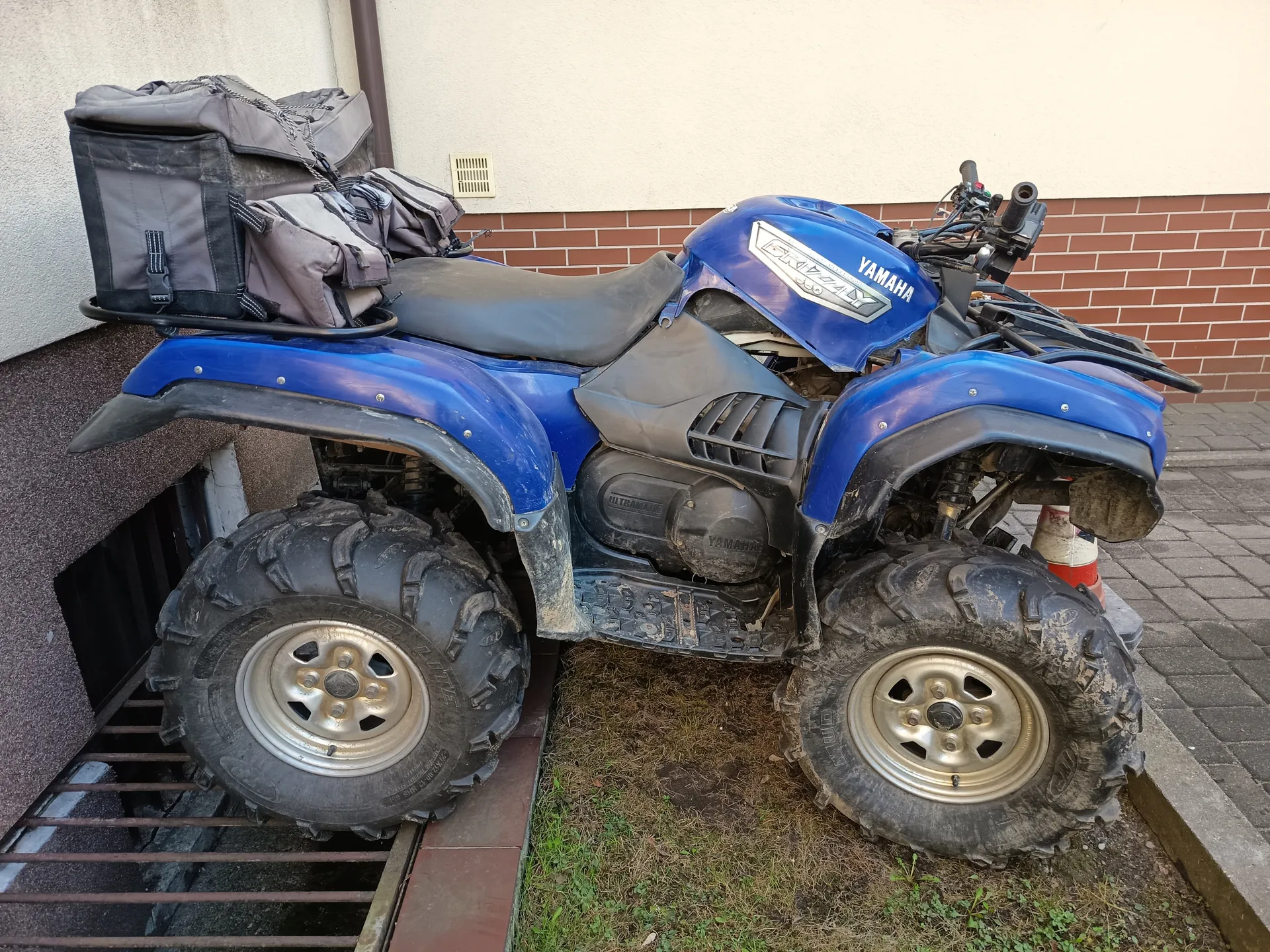 Niebieski quad Yamaha z czarnym siedzeniem i dużymi, brudnymi oponami. Na masce widoczny napis 'YAMAHA' oraz logo 'GRIZZLY'. Na tylnej części zamocowane są szare torby. Quad stoi przy budynku z ceglaną i jasną ścianą, obok metalowej kraty i pomarańczowo-białego klinu. W tle widoczna trawa i brukowana powierzchnia.