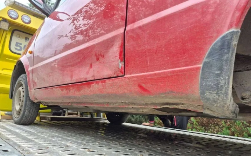 Zbliżenie na prawy bok czerwonego samochodu osobowego, który znajduje się na platformie lawety. Na drzwiach pasażera oraz dolnym progu widoczne są wgniecenia i zarysowania lakieru. W tle po lewej stronie widoczny jest fragment żółtego pojazdu z napisem '50'.