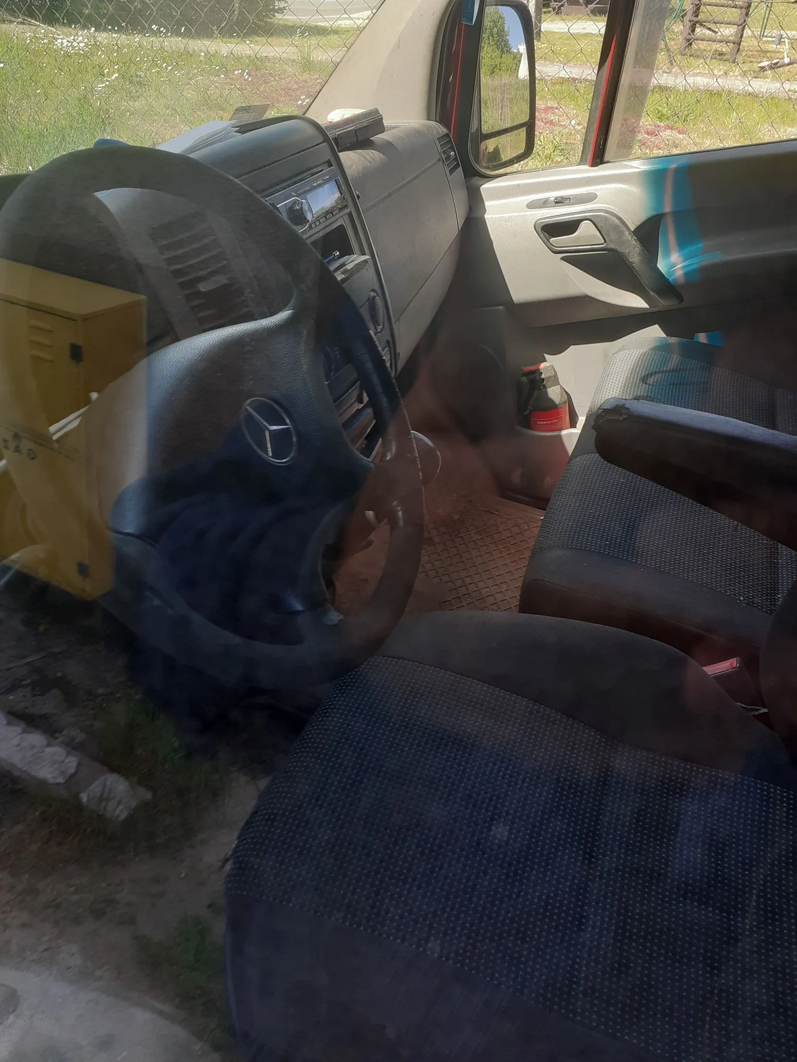 Wnętrze kabiny samochodu marki Mercedes-Benz widziane z perspektywy pasażera. Widoczna jest kierownica z logo marki, deska rozdzielcza z radiem, ciemna tapicerka foteli oraz drzwi pasażera. Zdjęcie zostało wykonane przez szybę, co powoduje widoczne odbicia.