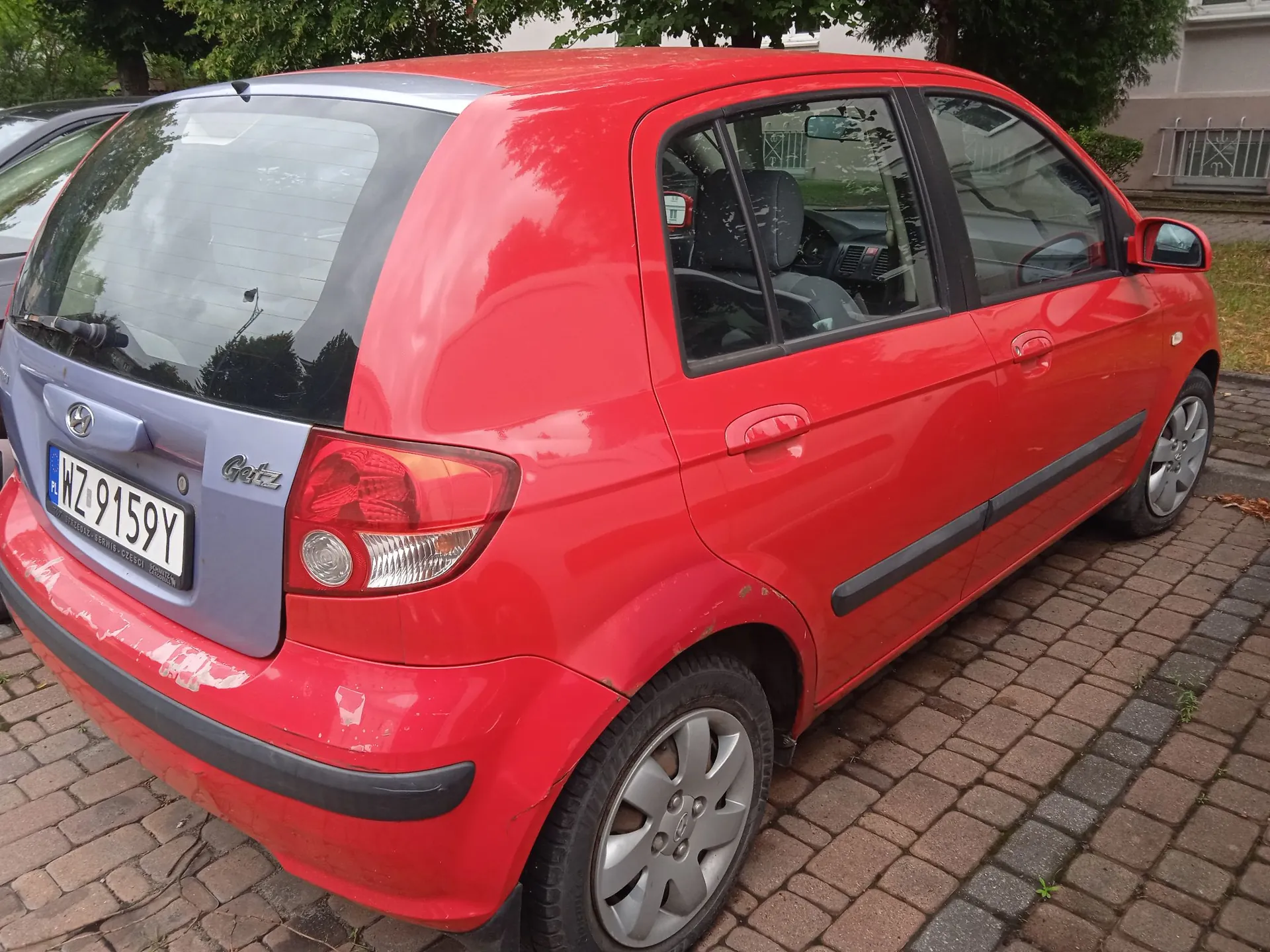 Czerwony samochód marki Hyundai Getz zaparkowany na kostce brukowej. Widoczny jest tył oraz prawy bok pojazdu. Samochód ma tablicę rejestracyjną o numerze WZ 9159Y. Na klapie bagażnika znajduje się napis 'Getz'.