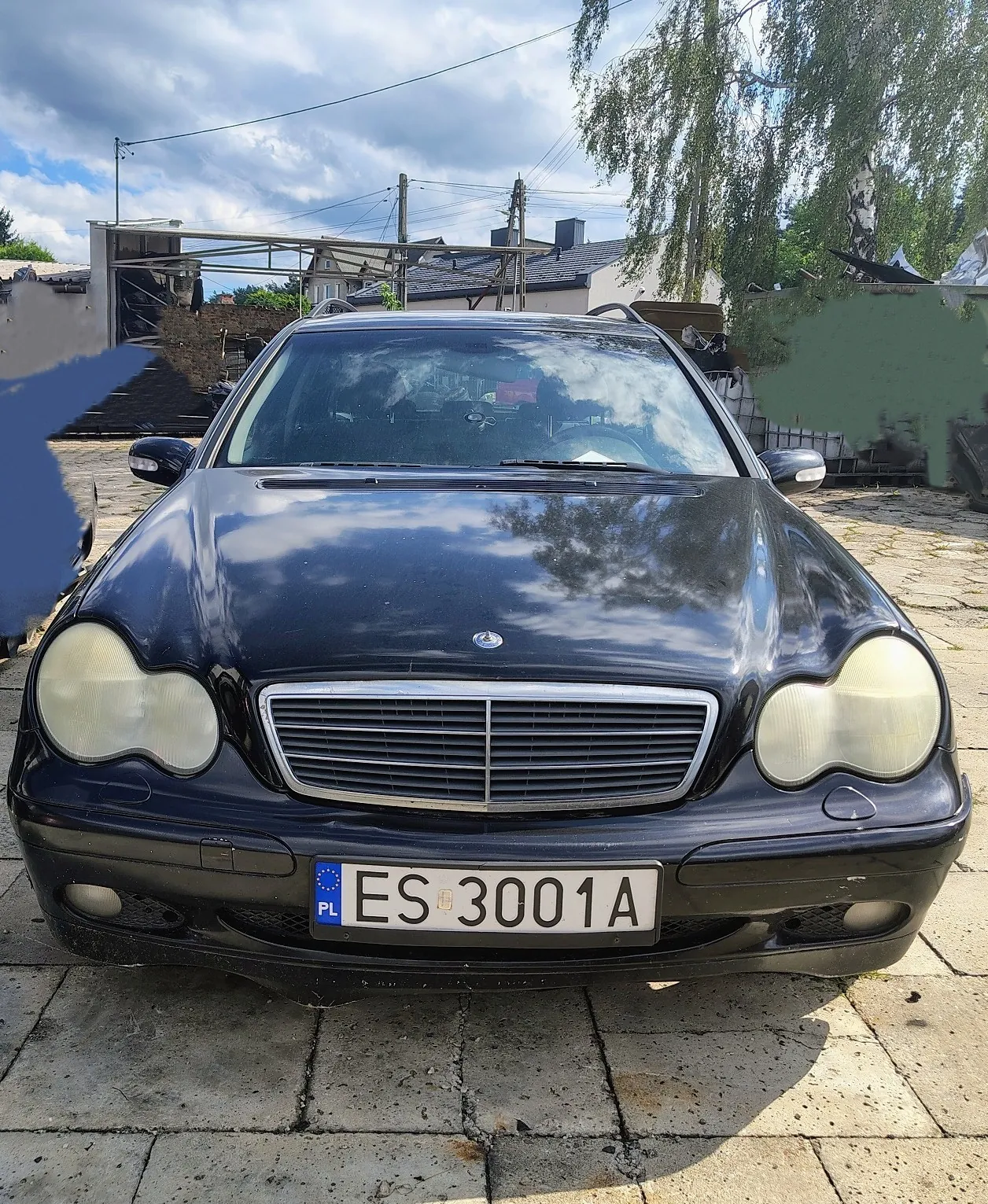 Czarny samochód marki Mercedes-Benz zaparkowany na kostce brukowej. Widok od przodu. Pojazd ma zmatowione reflektory i polską tablicę rejestracyjną o numerze ES 3001A. W tle widoczne są budynki, drzewa oraz zachmurzone niebo.