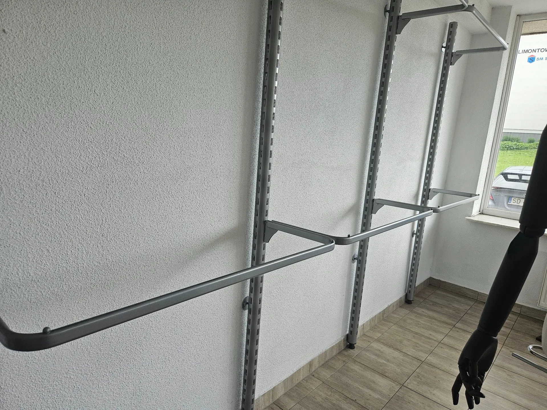 Metalowy system regałów ściennych zamontowany na białej, chropowatej ścianie. System składa się z pionowych szyn z otworami oraz poziomych rurek pełniących funkcję półek lub wieszaków. W tle widoczne jest okno z widokiem na zewnątrz oraz fragment czarnej rury lub elementu konstrukcyjnego po prawej stronie. Podłoga wyłożona jest jasnymi płytkami.