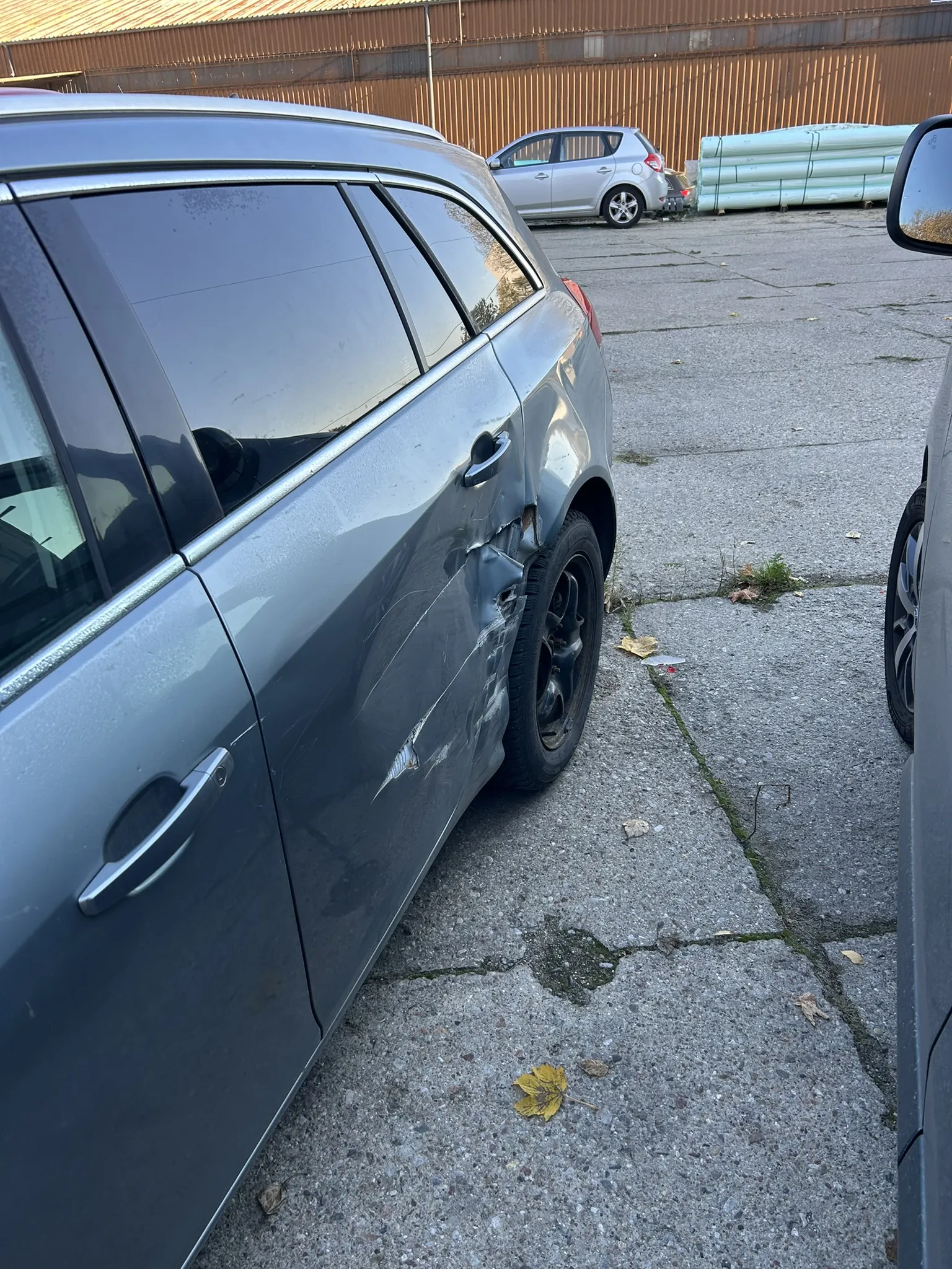 Zdjęcie przedstawia szary samochód typu kombi z widocznymi uszkodzeniami na tylnej prawej stronie nadwozia. Pojazd stoi na betonowej powierzchni z pęknięciami, wśród których rosną niewielkie rośliny i leżą opadłe liście. W tle widoczny jest srebrny samochód osobowy oraz stos białych rur. Za nimi znajduje się brązowa metalowa ściana budynku przemysłowego. Po prawej stronie kadru widoczna jest część innego pojazdu z lusterkiem bocznym.