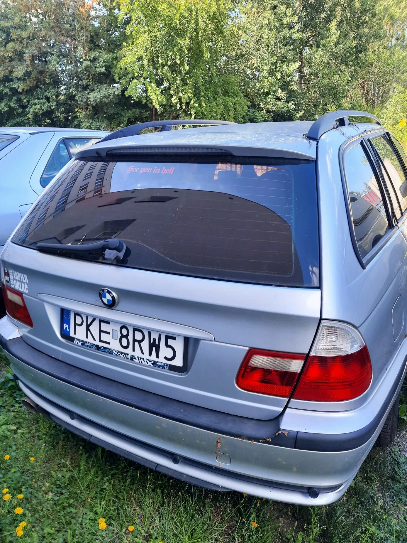 Tylny widok srebrnego samochodu marki BMW. Na tylnej szybie znajduje się czerwony napis „See you in hell”. Samochód ma polską tablicę rejestracyjną o numerze PKE 8RW5. Pojazd stoi na trawie, w tle widoczne są zielone drzewa.